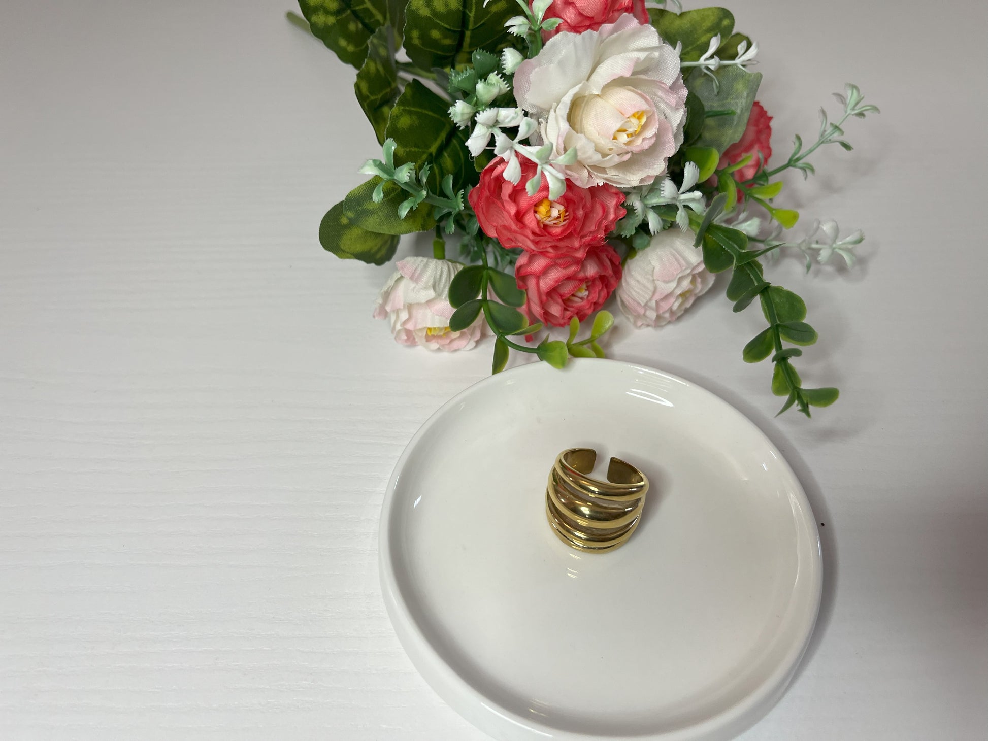 Anello Fascia Tripla placcato oro con tre fasce sovrapposte eleganti, design moderno e minimalista su piattino bianco con fiore rosa e bianco