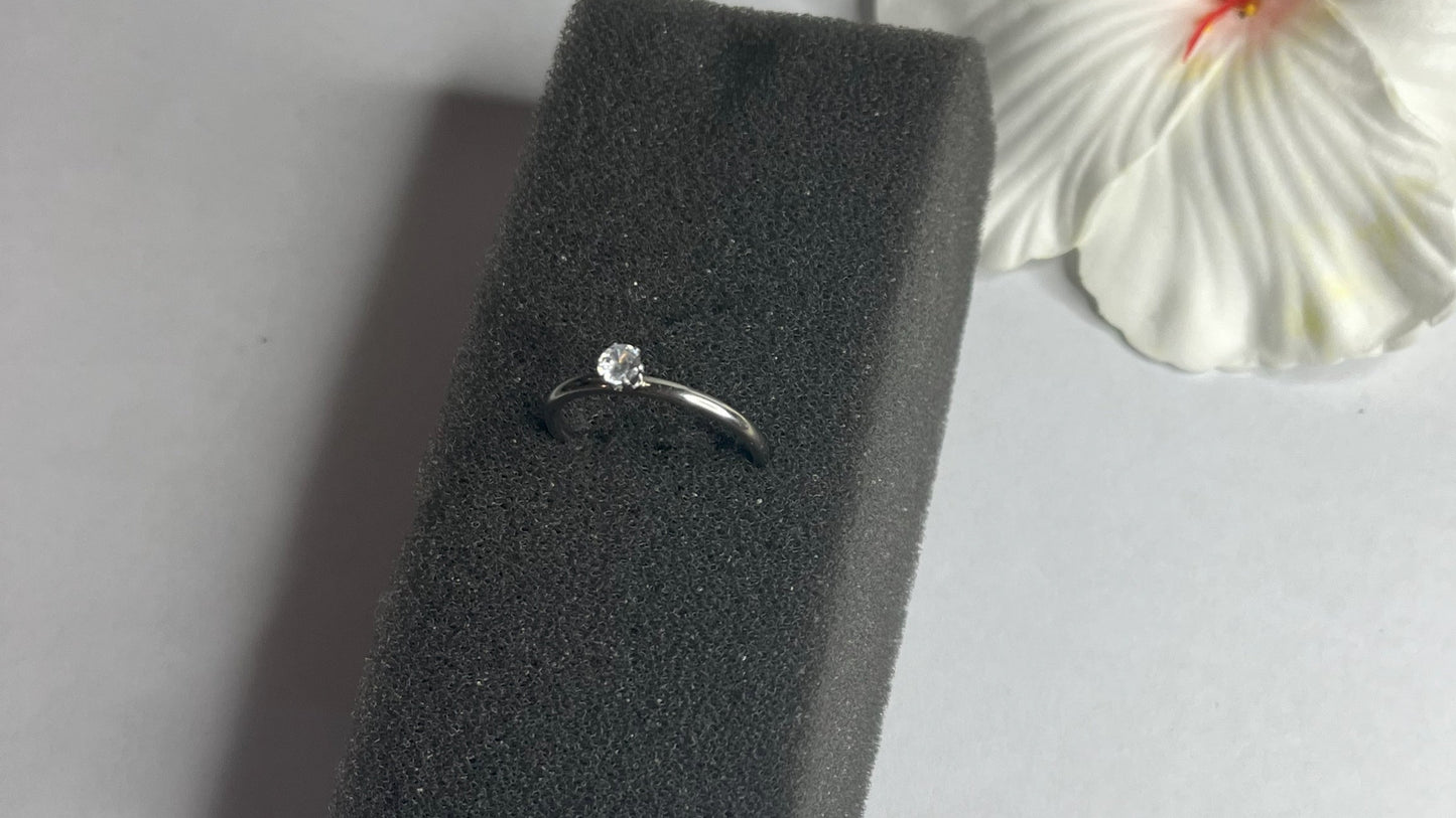 Anello solitario in acciaio placcato argento con pietra brillante centrale, montatura classica elegante su superficie grigia con conchiglia decorativa