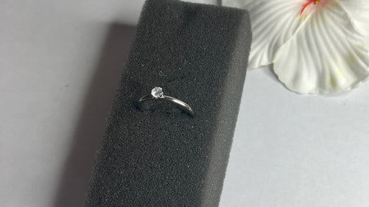 Anello solitario in acciaio placcato argento con pietra brillante centrale, montatura classica elegante su superficie grigia con conchiglia decorativa