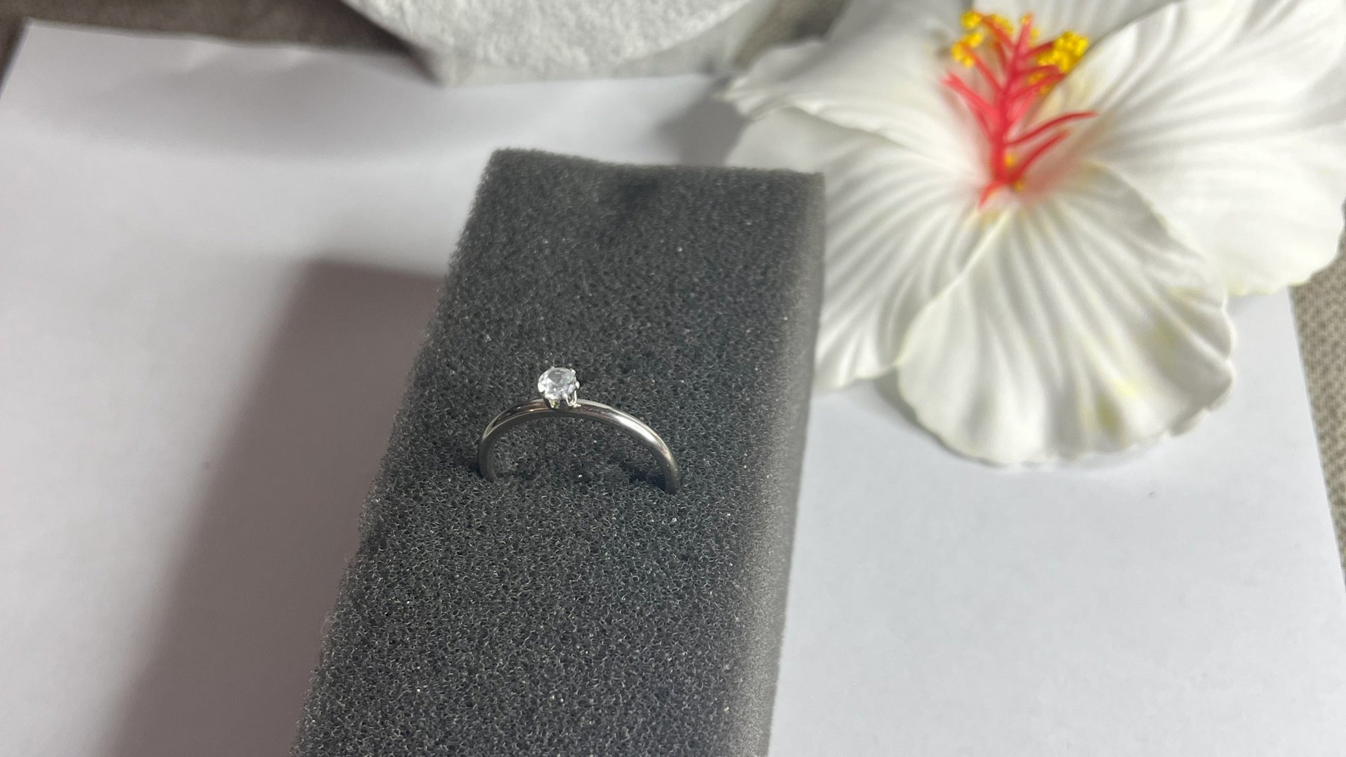 Anello solitario in acciaio placcato argento con pietra brillante centrale, montatura classica elegante su superficie grigia con conchiglia decorativa