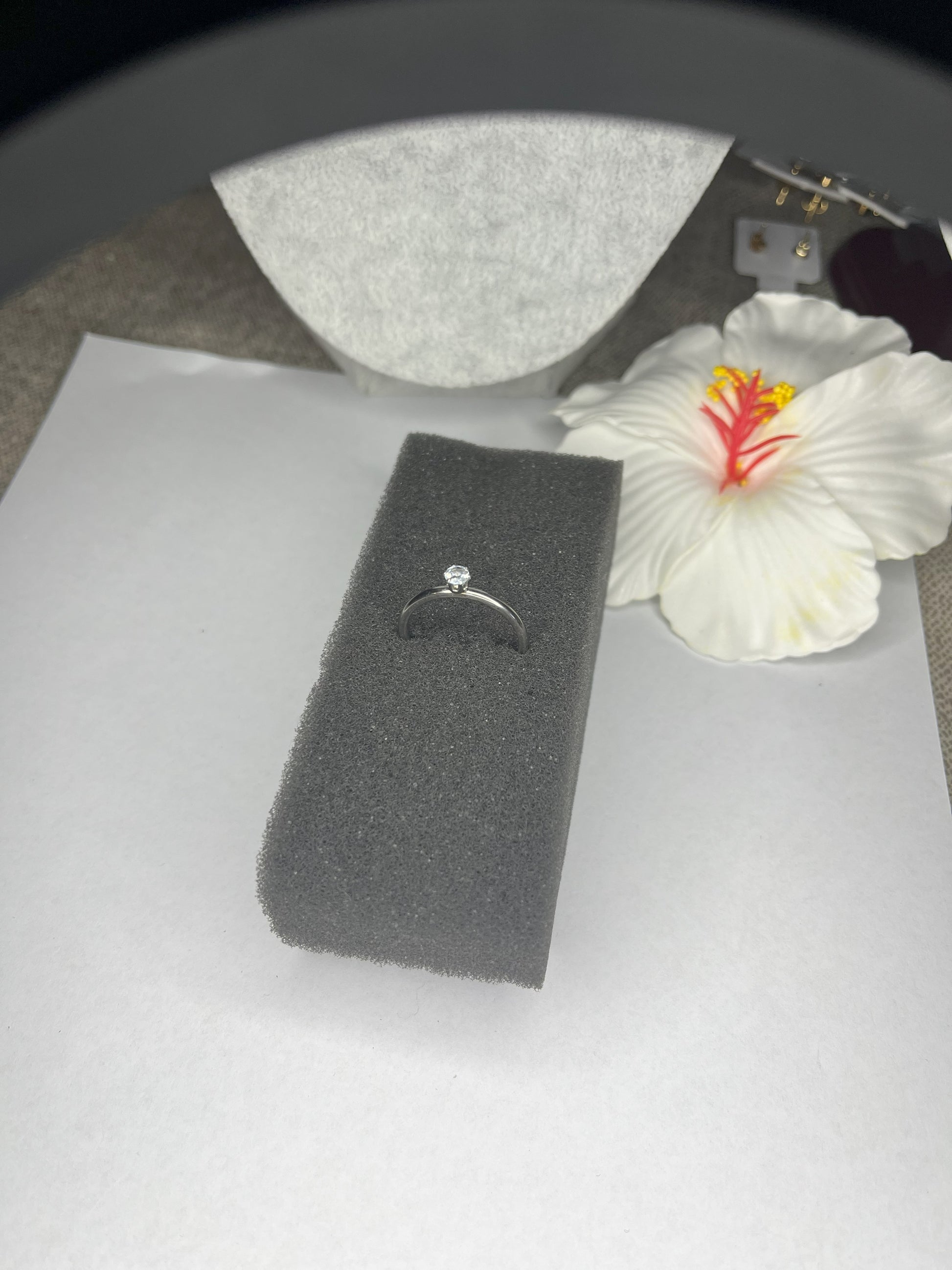 Anello solitario in acciaio placcato argento con pietra brillante centrale, montatura classica elegante su superficie grigia con conchiglia decorativa