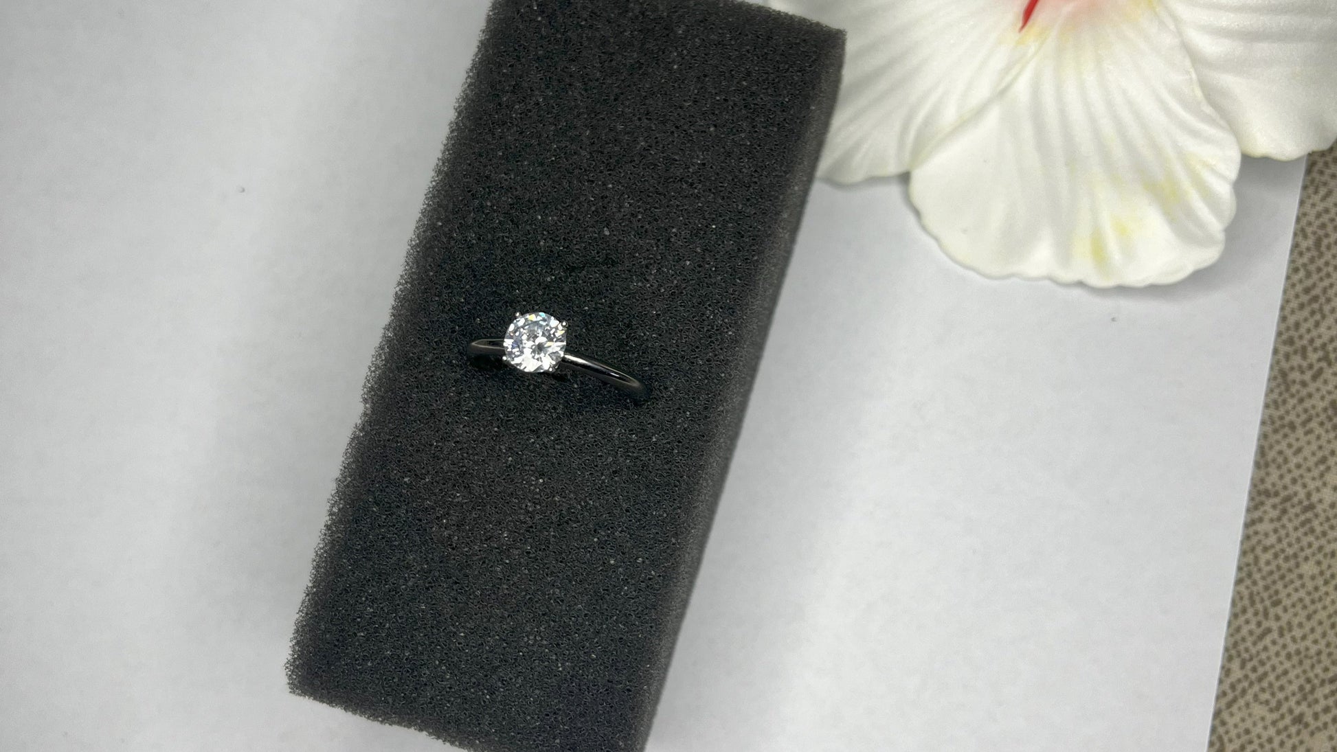 Anello solitario in acciaio placcato argento con pietra brillante centrale, montatura classica elegante su superficie grigia con conchiglia decorativa