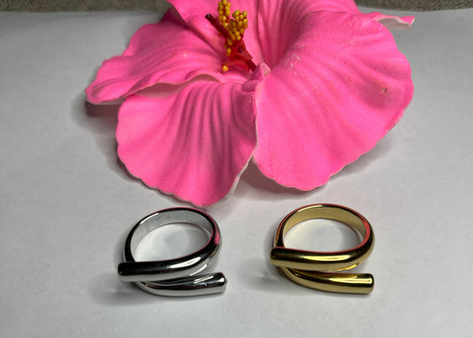 Anello Twist in acciaio premium con design avvolgente moderno, disponibile in placcatura oro e argento, fotografato su sfondo neutro con petali rosa per evidenziare l'eleganza contemporanea