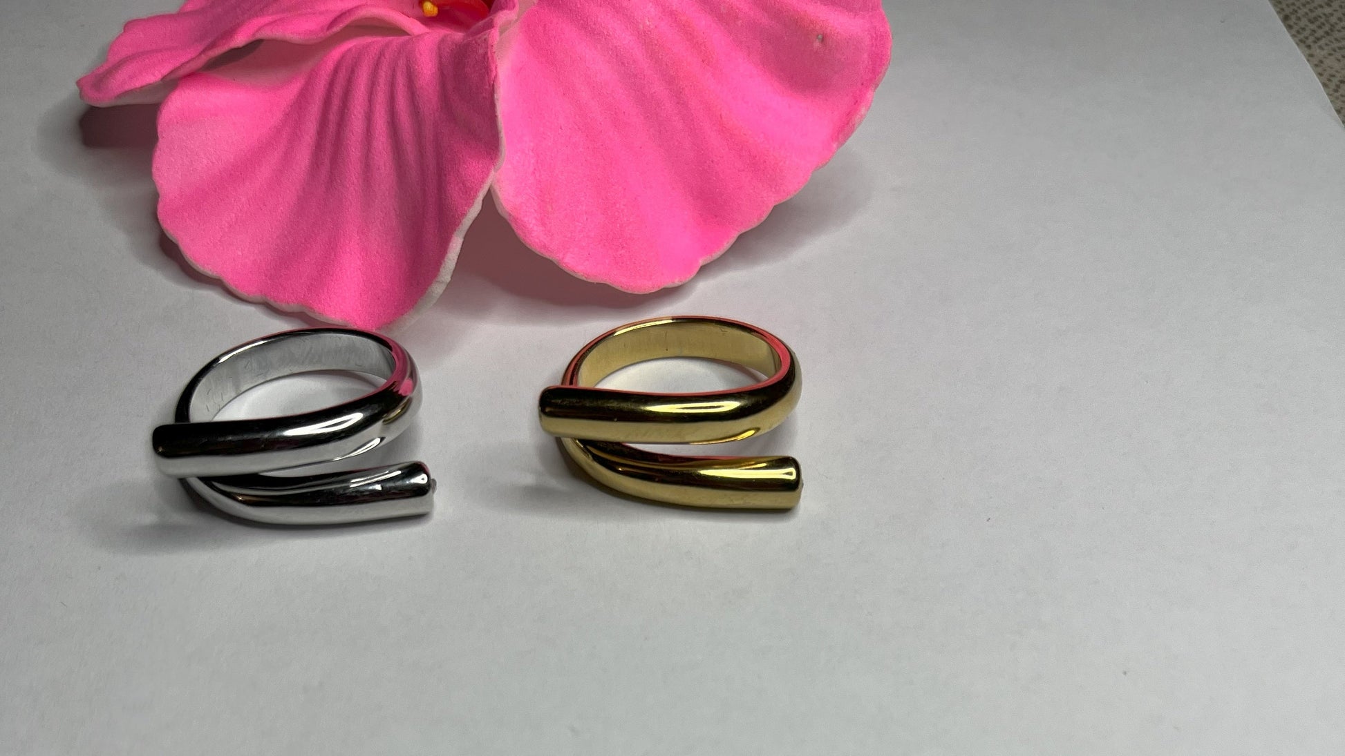 Anello Twist in acciaio premium con design avvolgente moderno, disponibile in placcatura oro e argento, fotografato su sfondo neutro con petali rosa per evidenziare l'eleganza contemporanea