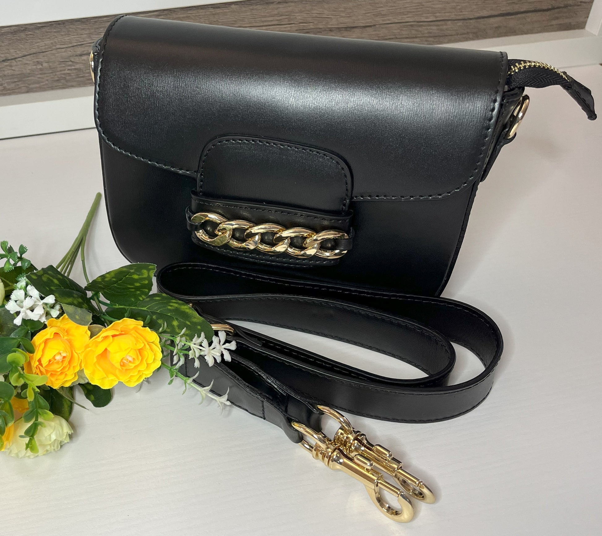 Borsa Toscana Chic in vera pelle nera Made in Italy con catena dorata decorativa e tracolla regolabile, fotografata su superficie bianca con fiori gialli