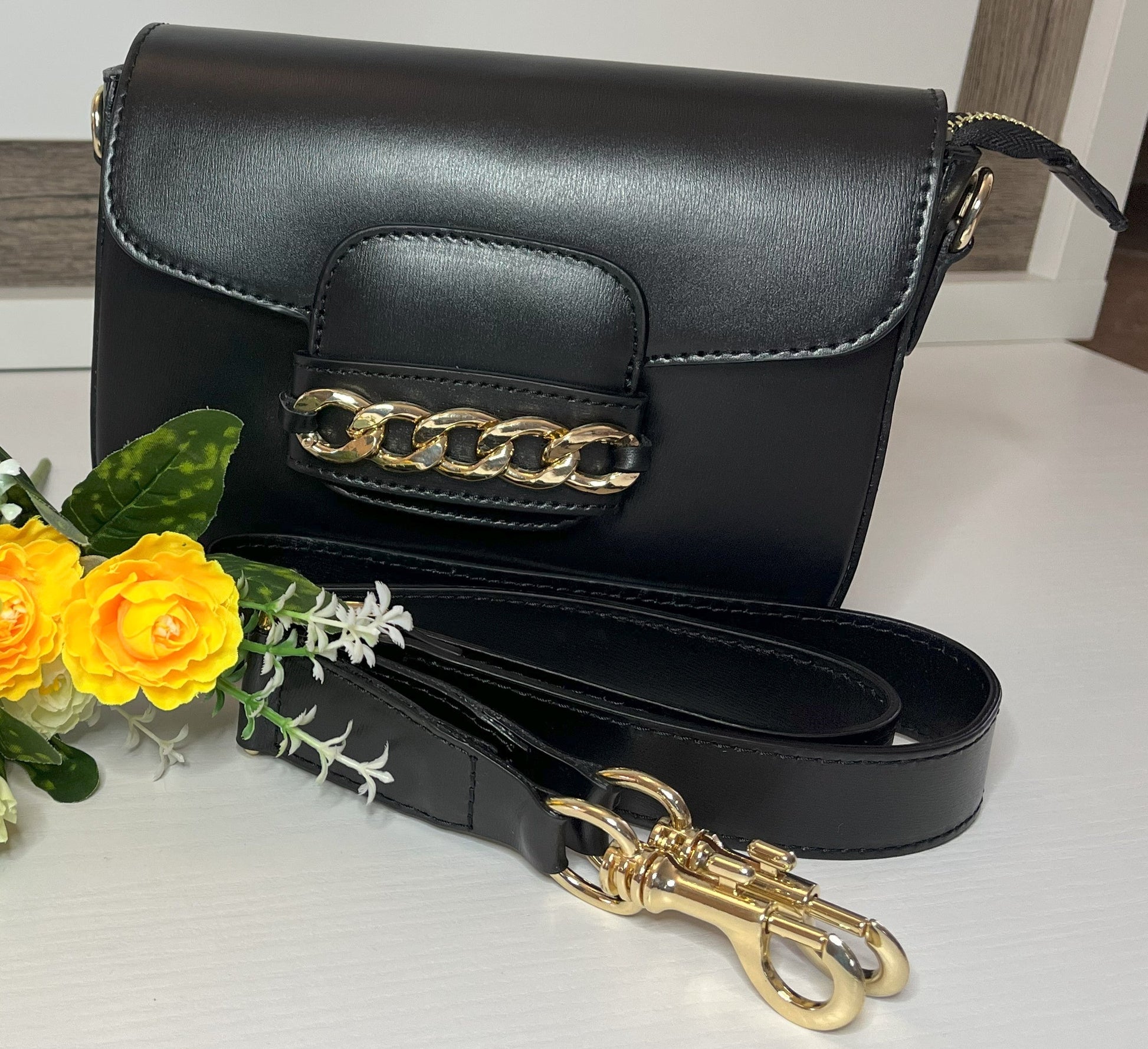 Borsa Toscana Chic in vera pelle nera Made in Italy con catena dorata decorativa e tracolla regolabile, fotografata su superficie bianca con fiori gialli