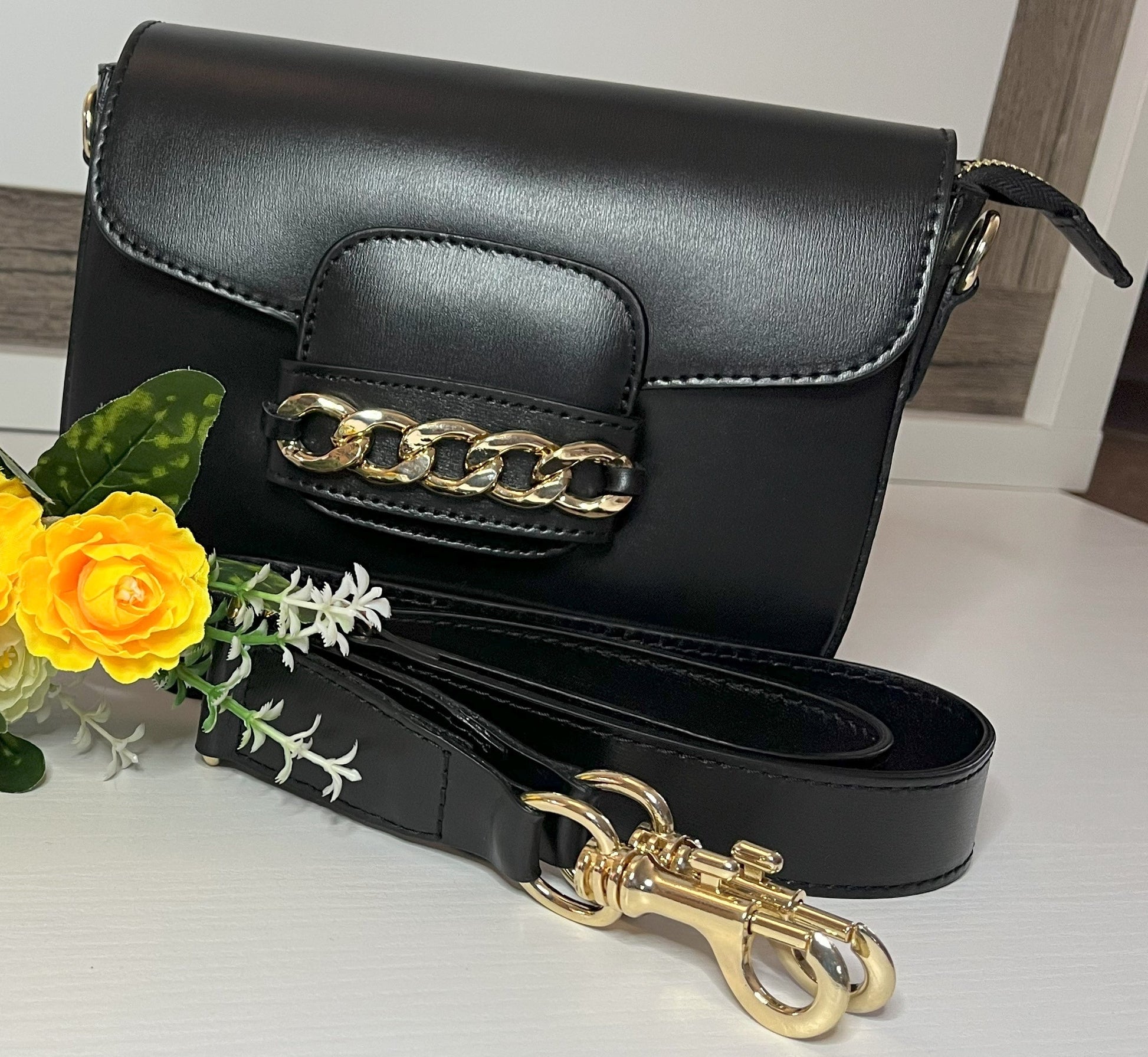 Borsa Toscana Chic in vera pelle nera Made in Italy con catena dorata decorativa e tracolla regolabile, fotografata su superficie bianca con fiori gialli