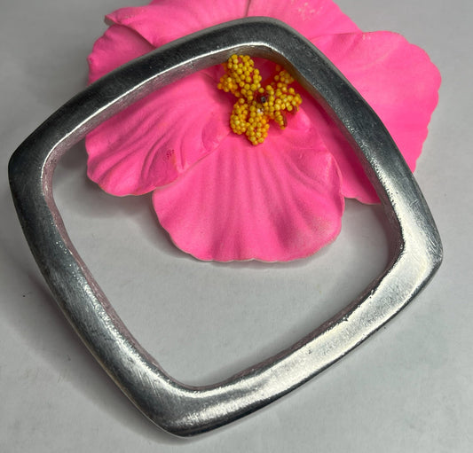 Bracciale quadro in alluminio con design geometrico moderno e finitura satinata, fotografato accanto a fiore rosa