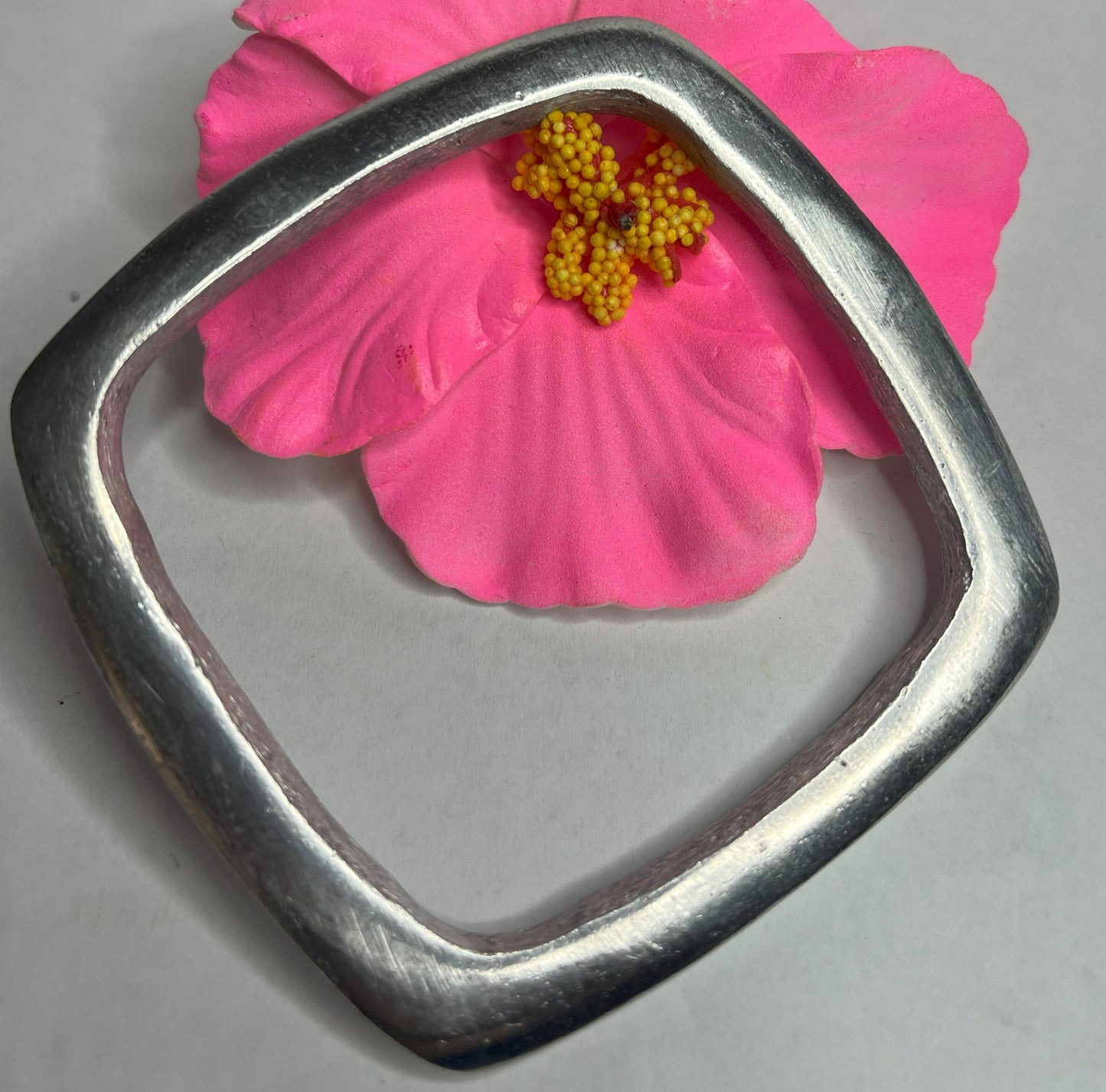 Bracciale quadro in alluminio con design geometrico moderno e finitura satinata, fotografato accanto a fiore rosa