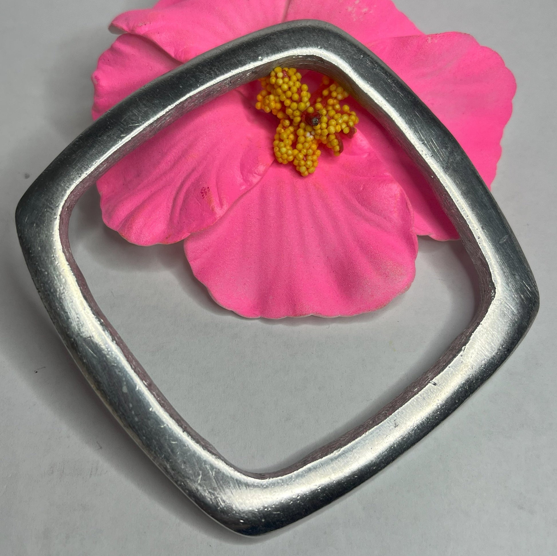 Bracciale quadro in alluminio con design geometrico moderno e finitura satinata, fotografato accanto a fiore rosa
