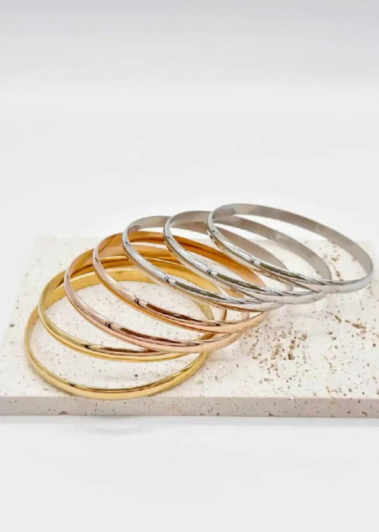Set completo di 7 braccialetti eleganti a sezione semicerchio in diverse tonalità di oro - oro giallo, oro rosa e oro bianco - disposti artisticamente su superficie bianca con texture naturale. Design minimalista e confortevole perfetto per layering.