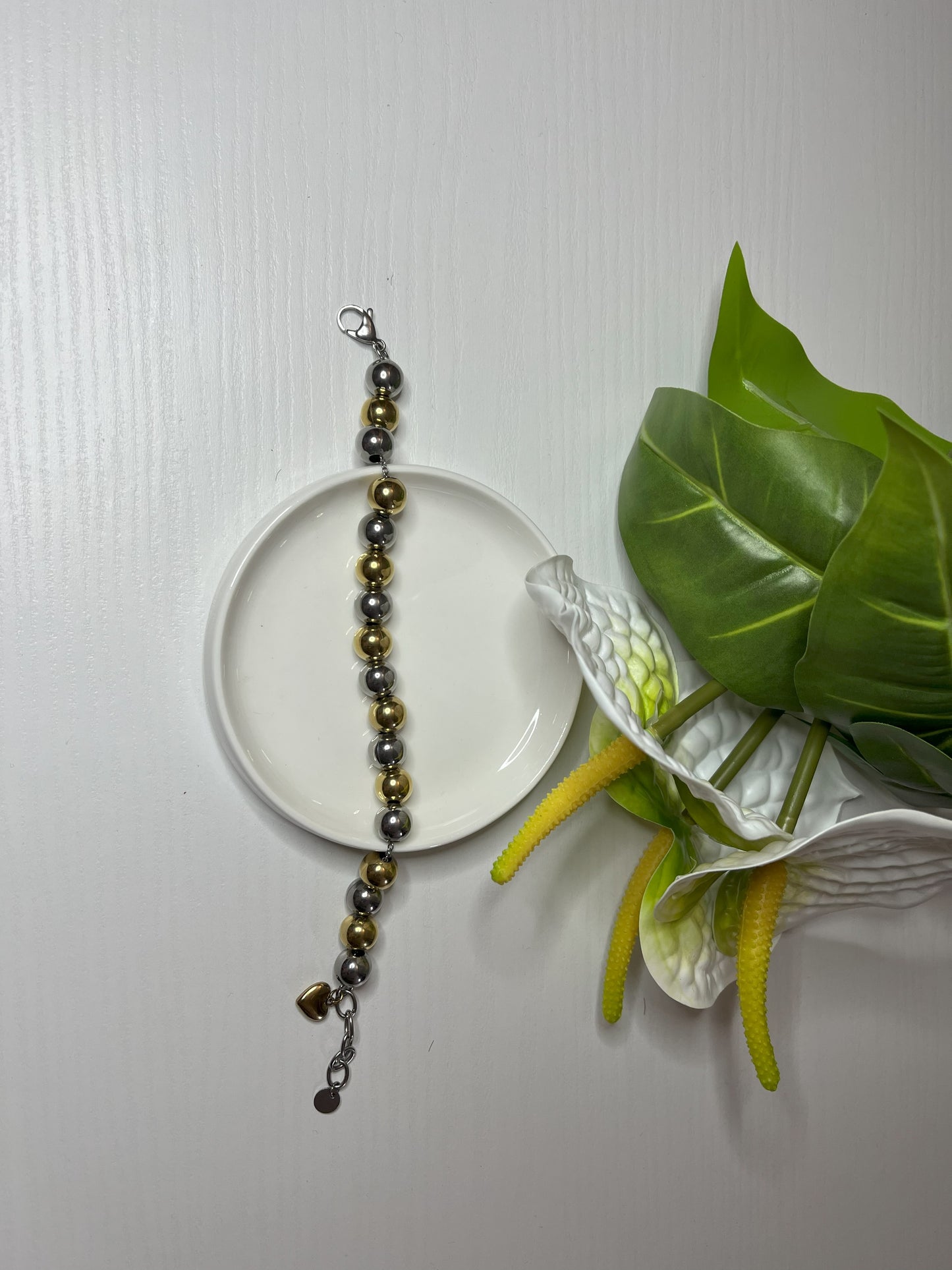 Elegante braccialetto con sfere dorate e argentate lucide su piattino bianco, decorato con fiori bianchi e foglie verdi