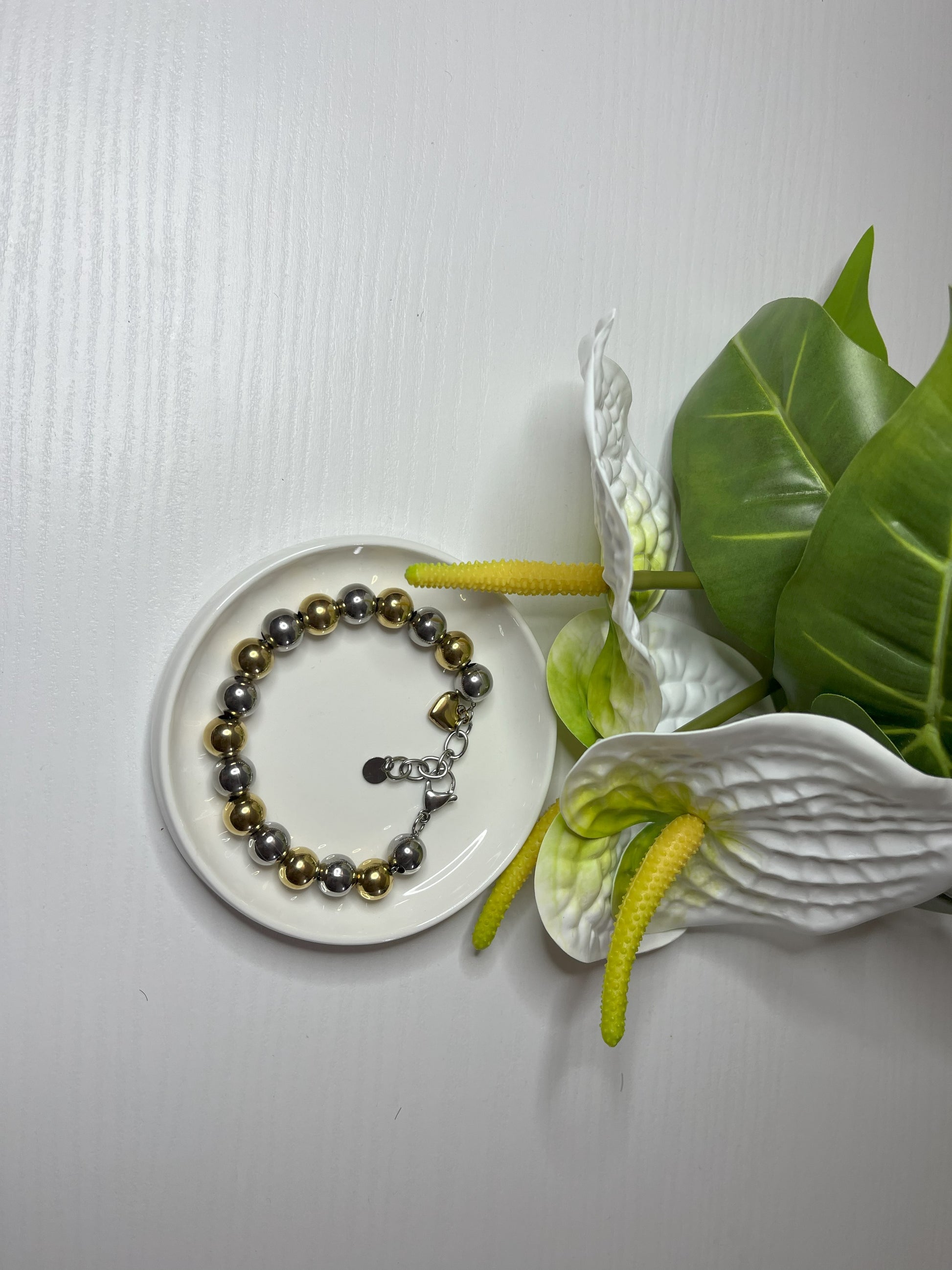 Elegante braccialetto con sfere dorate e argentate lucide su piattino bianco, decorato con fiori bianchi e foglie verdi