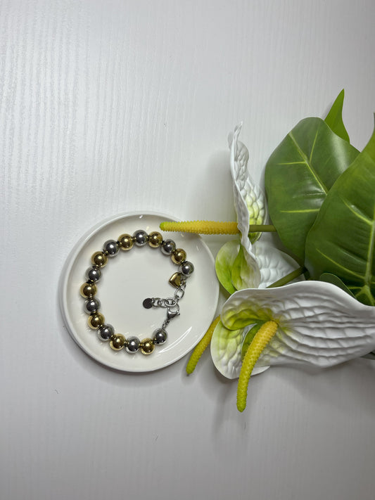Elegante braccialetto con sfere dorate e argentate lucide su piattino bianco, decorato con fiori bianchi e foglie verdi