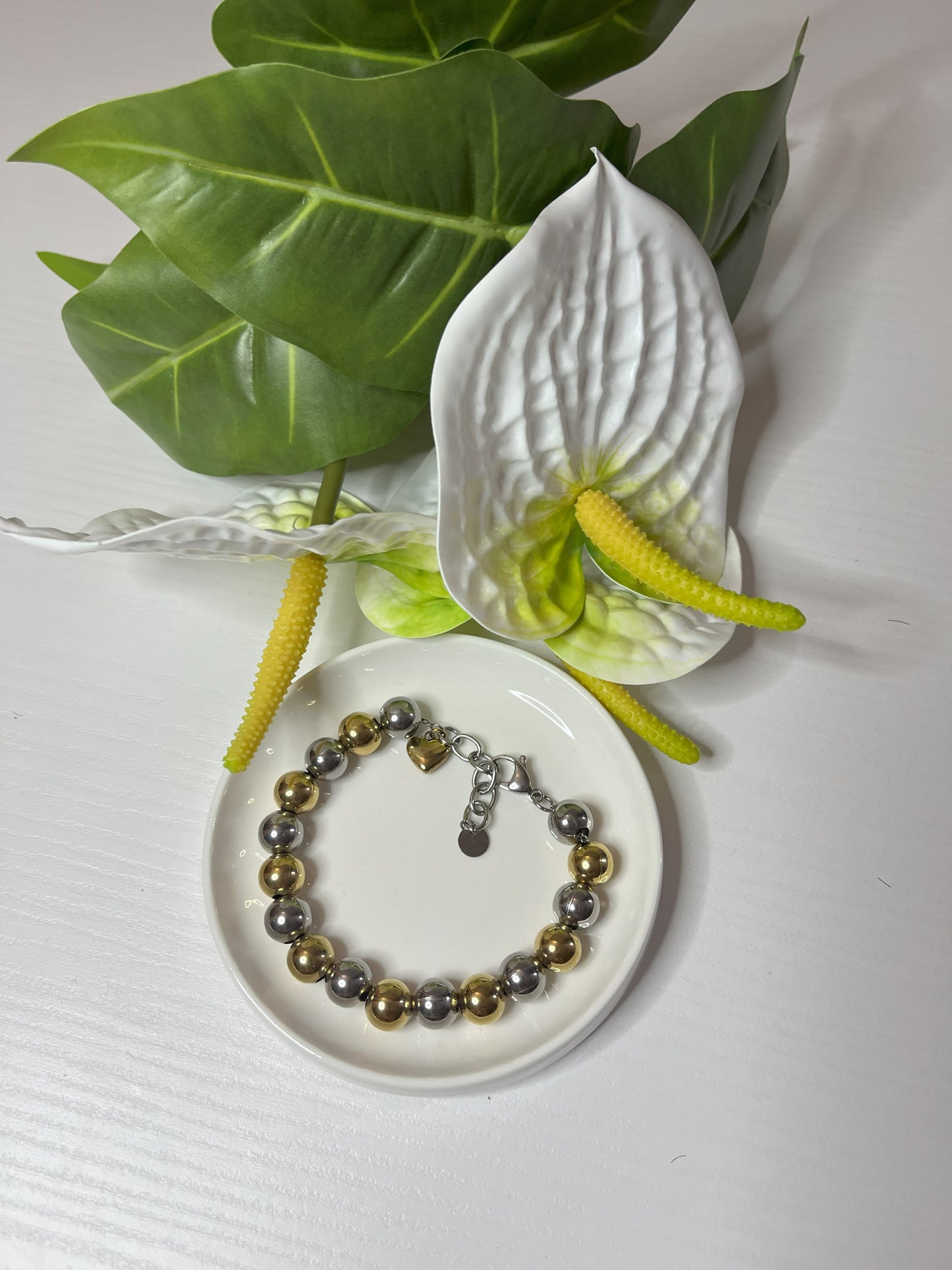 Elegante braccialetto con sfere dorate e argentate lucide su piattino bianco, decorato con fiori bianchi e foglie verdi