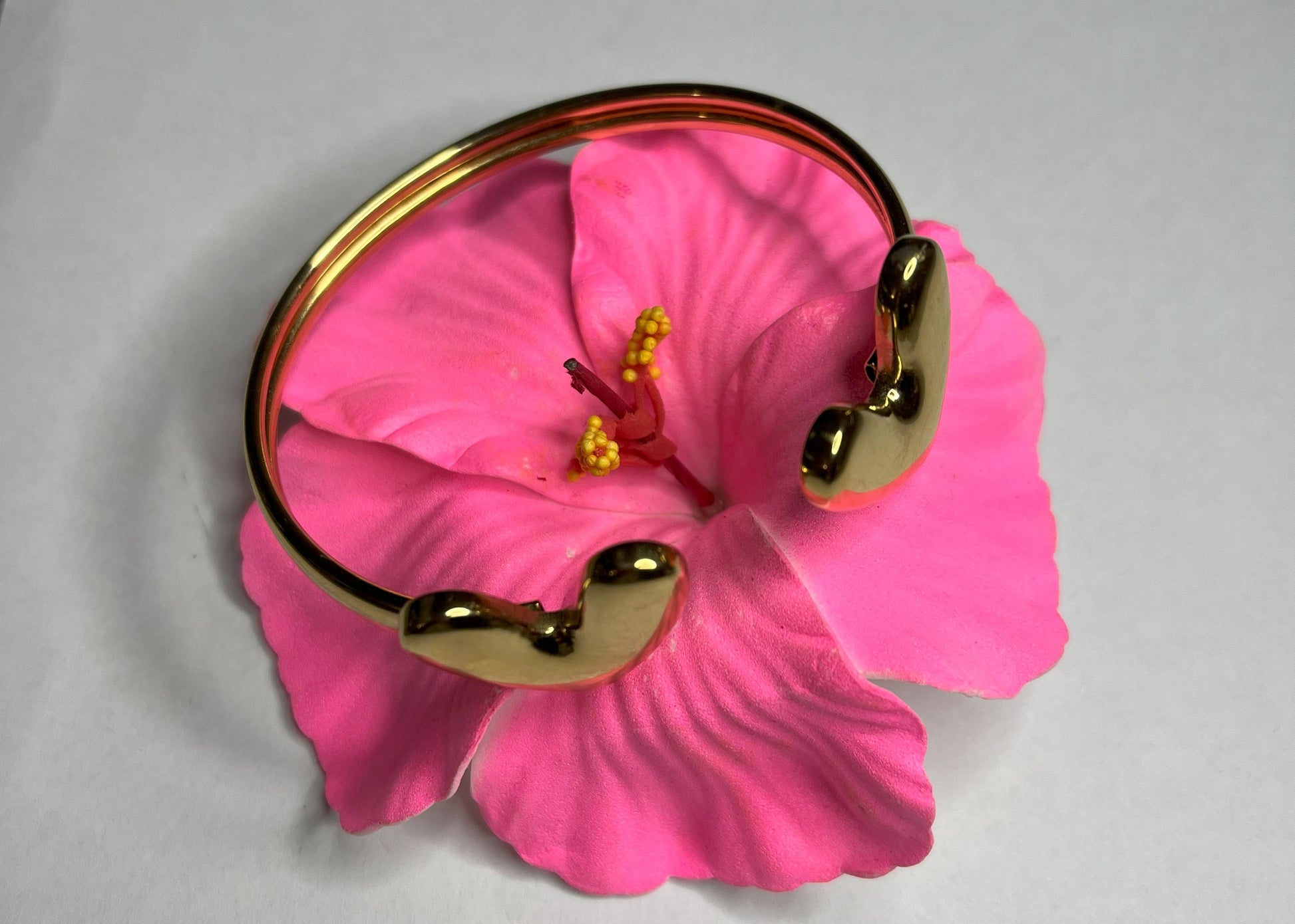 Braccialetto Due Cuori in acciaio premium placcato oro con design aperto regolabile, fotografato su fiore rosa fucsia per evidenziare l'eleganza romantica del gioiello