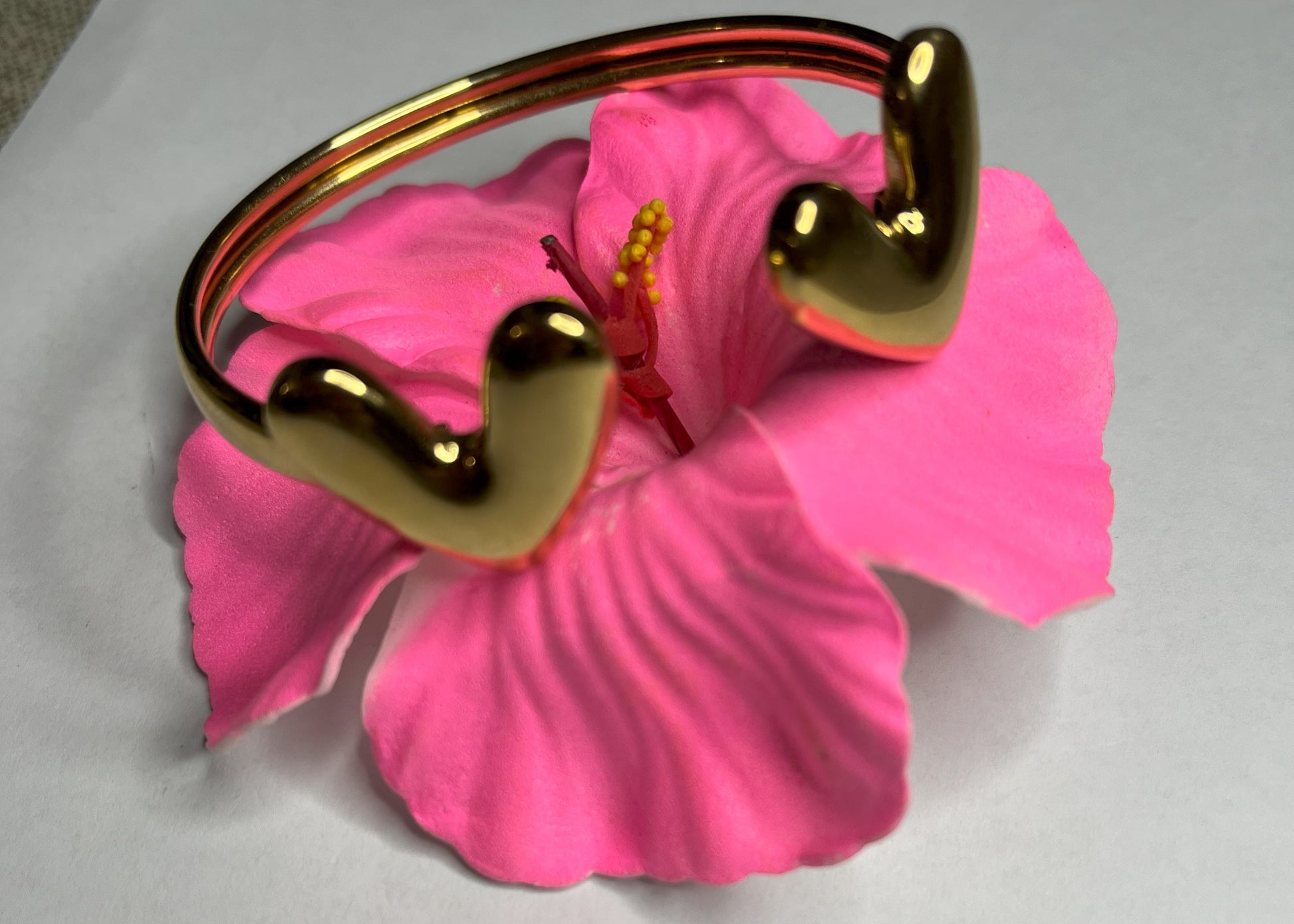 Braccialetto Due Cuori in acciaio premium placcato oro con design aperto regolabile, fotografato su fiore rosa fucsia per evidenziare l'eleganza romantica del gioiello