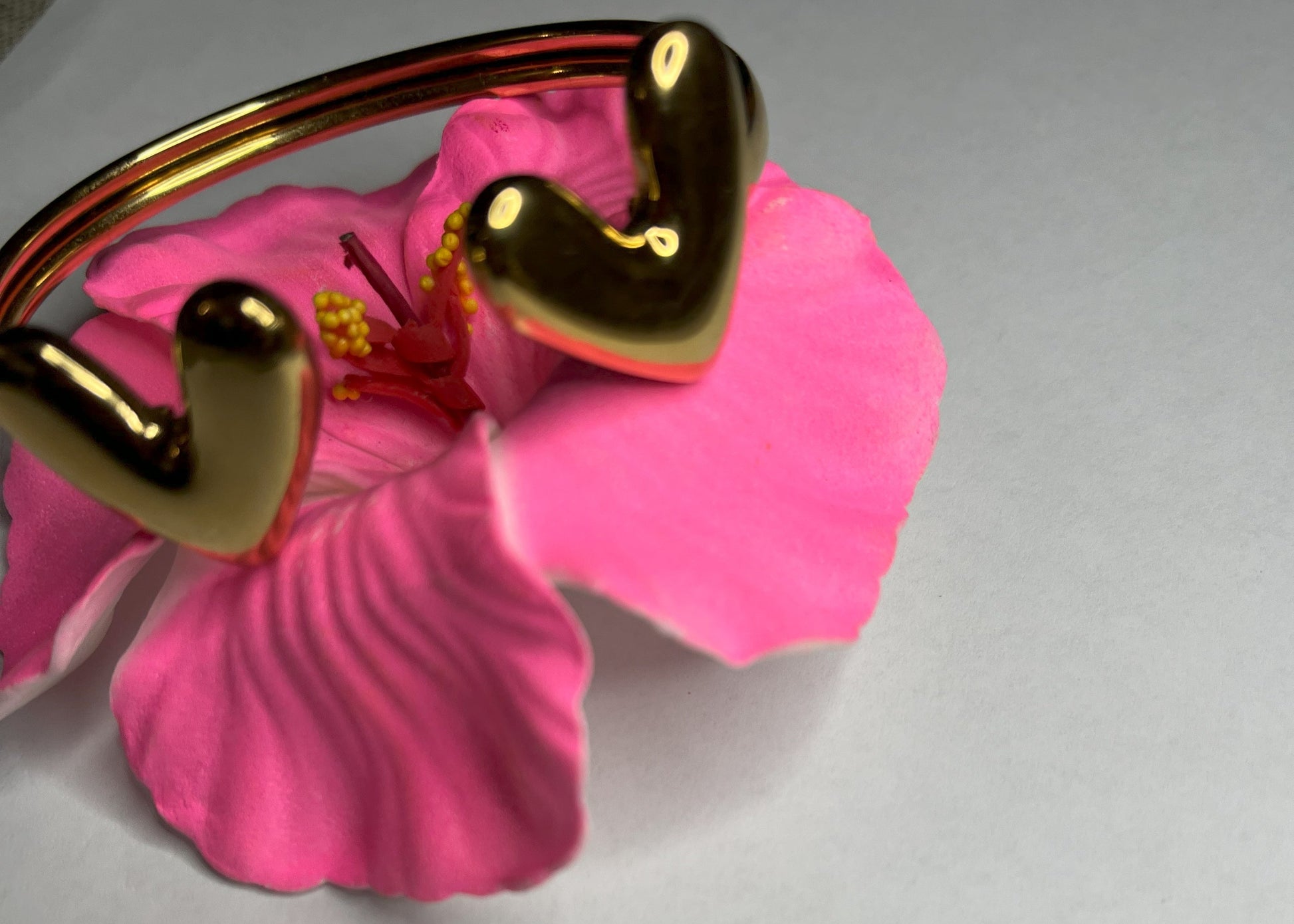 Braccialetto Due Cuori in acciaio premium placcato oro con design aperto regolabile, fotografato su fiore rosa fucsia per evidenziare l'eleganza romantica del gioiello