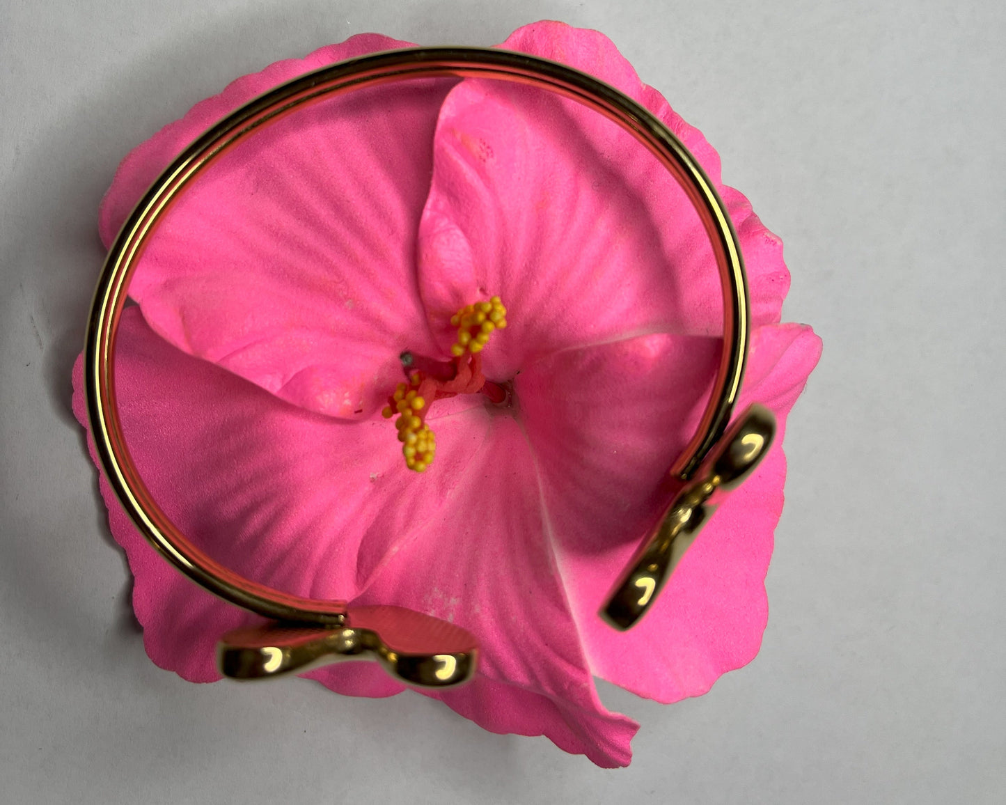 Braccialetto Due Cuori in acciaio premium placcato oro con design aperto regolabile, fotografato su fiore rosa fucsia per evidenziare l'eleganza romantica del gioiello