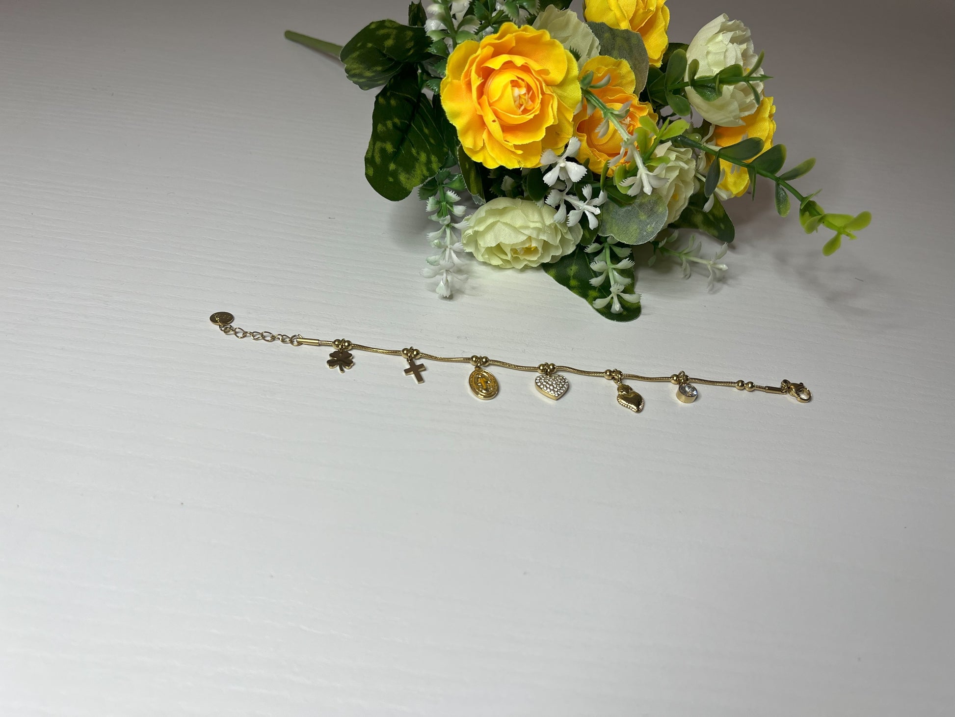 Braccialetto Zoe con charm dorati inclusi croce, medaglia religiosa, cuore brillante e cristallo su piatto bianco con fiori gialli