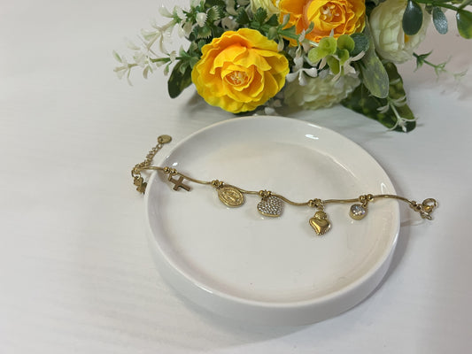 Braccialetto Zoe con charm dorati inclusi croce, medaglia religiosa, cuore brillante e cristallo su piatto bianco con fiori gialli