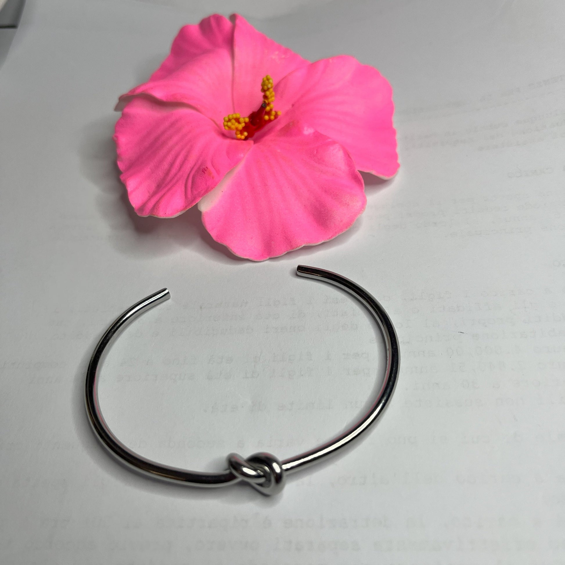 Braccialetto Nodo d'Amore Rigido in acciaio premium placcato argento con nodo centrale simbolico, accanto a petali di rosa fucsia su sfondo grigio