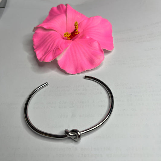 Braccialetto Nodo d'Amore Rigido in acciaio premium placcato argento con nodo centrale simbolico, accanto a petali di rosa fucsia su sfondo grigio