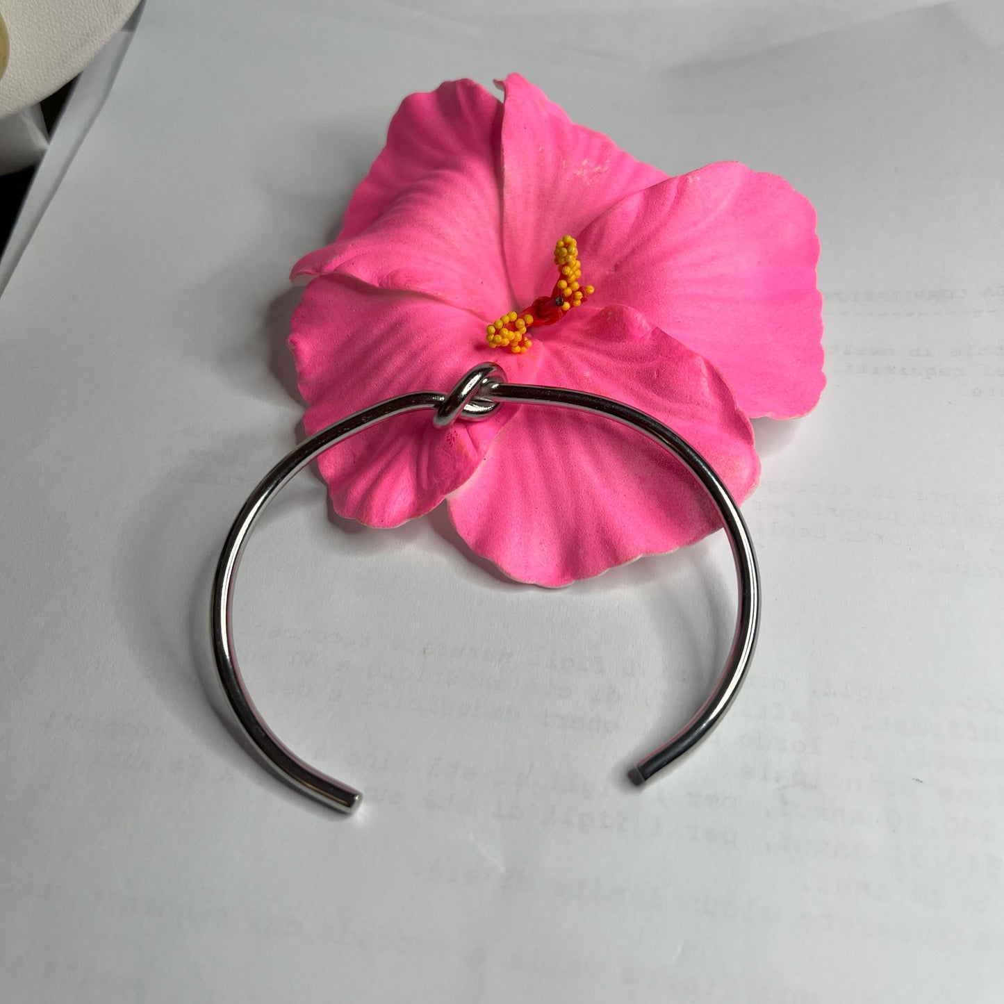 Braccialetto Nodo d'Amore Rigido in acciaio premium placcato argento con nodo centrale simbolico, accanto a petali di rosa fucsia su sfondo grigio