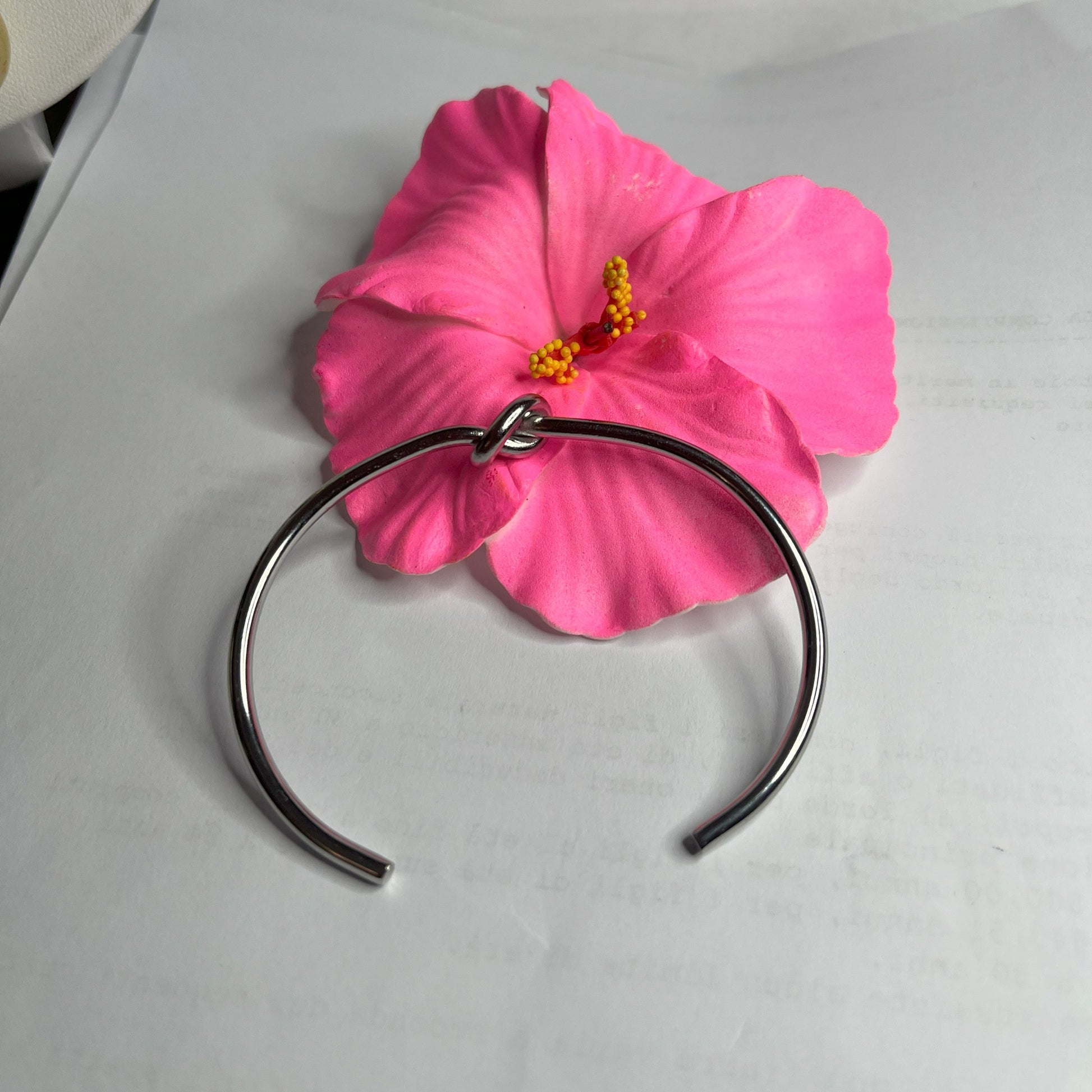 Braccialetto Nodo d'Amore Rigido in acciaio premium placcato argento con nodo centrale simbolico, accanto a petali di rosa fucsia su sfondo grigio