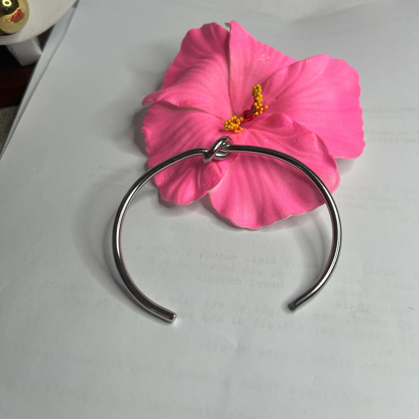 Braccialetto Nodo d'Amore Rigido in acciaio premium placcato argento con nodo centrale simbolico, accanto a petali di rosa fucsia su fondo grigio