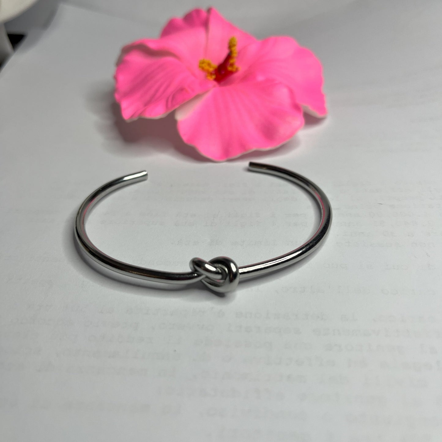 Braccialetto Nodo d'Amore Rigido in acciaio premium placcato argento con nodo centrale simbolico, accanto a petali di rosa fucsia su sfondo grigio