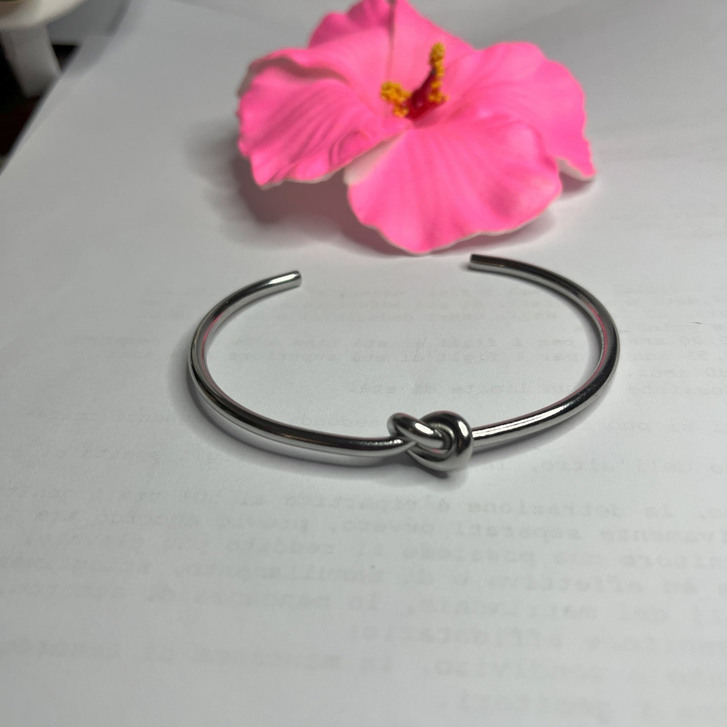 Braccialetto Nodo d'Amore Rigido in acciaio premium placcato argento con nodo centrale simbolico, accanto a petali di rosa fucsia su sfondo grigio
