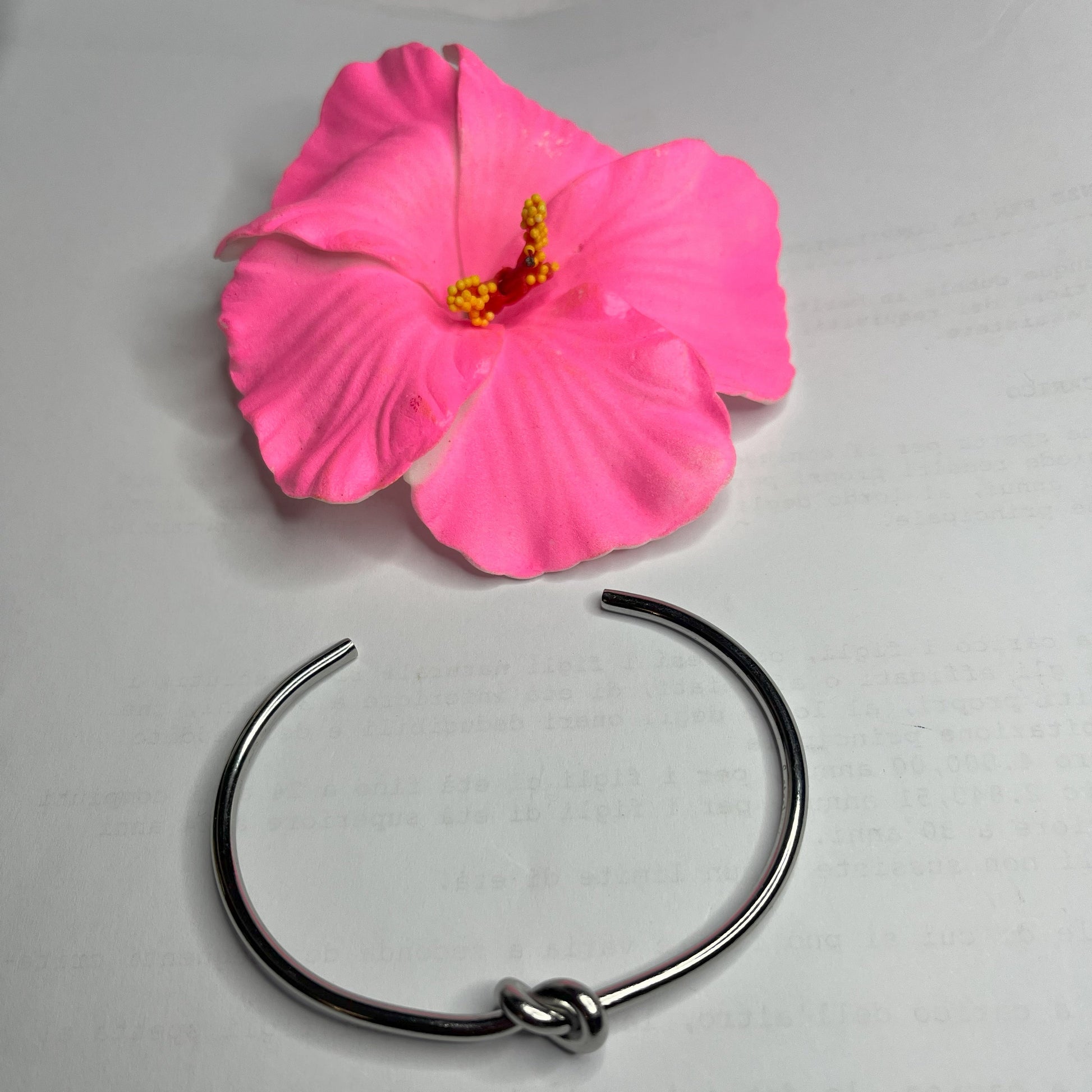 Braccialetto Nodo d'Amore Rigido in acciaio premium placcato argento con nodo centrale simbolico, accanto a petali di rosa fucsia su sfondo grigio
