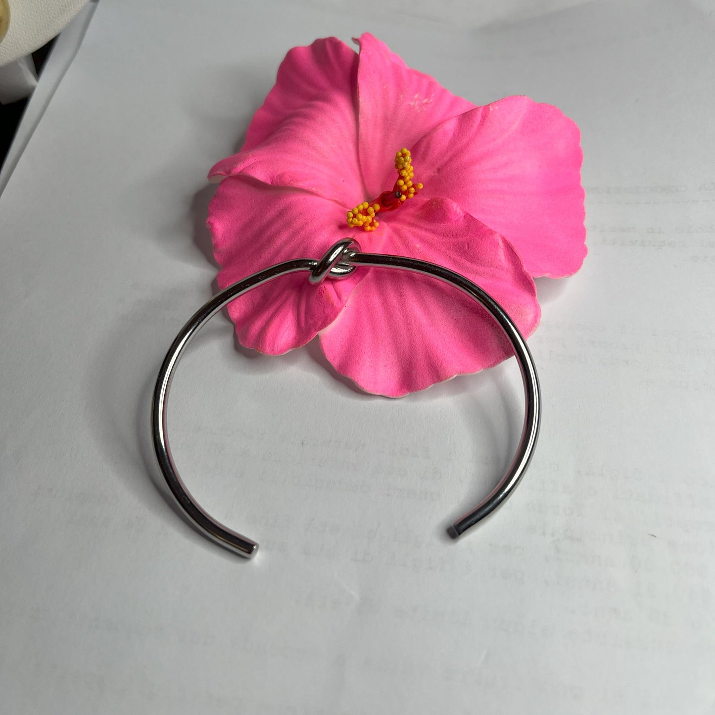Braccialetto Nodo d'Amore Rigido in acciaio premium placcato argento con nodo centrale simbolico, accanto a petali di rosa fucsia su sfondo grigio