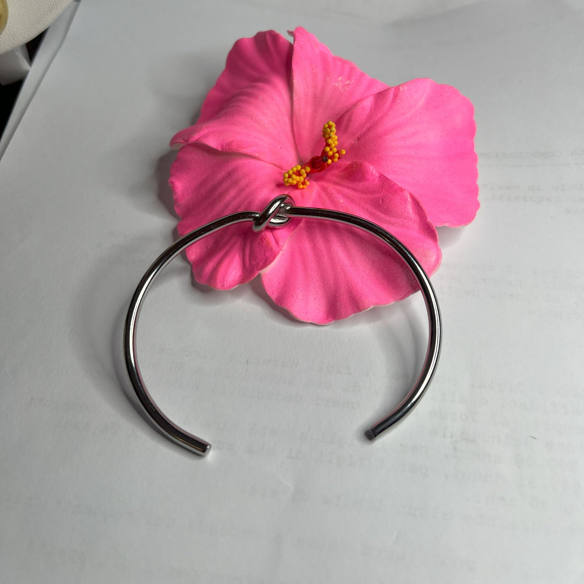 Braccialetto Nodo d'Amore Rigido in acciaio premium placcato argento con nodo centrale simbolico, accanto a petali di rosa fucsia su sfondo grigio