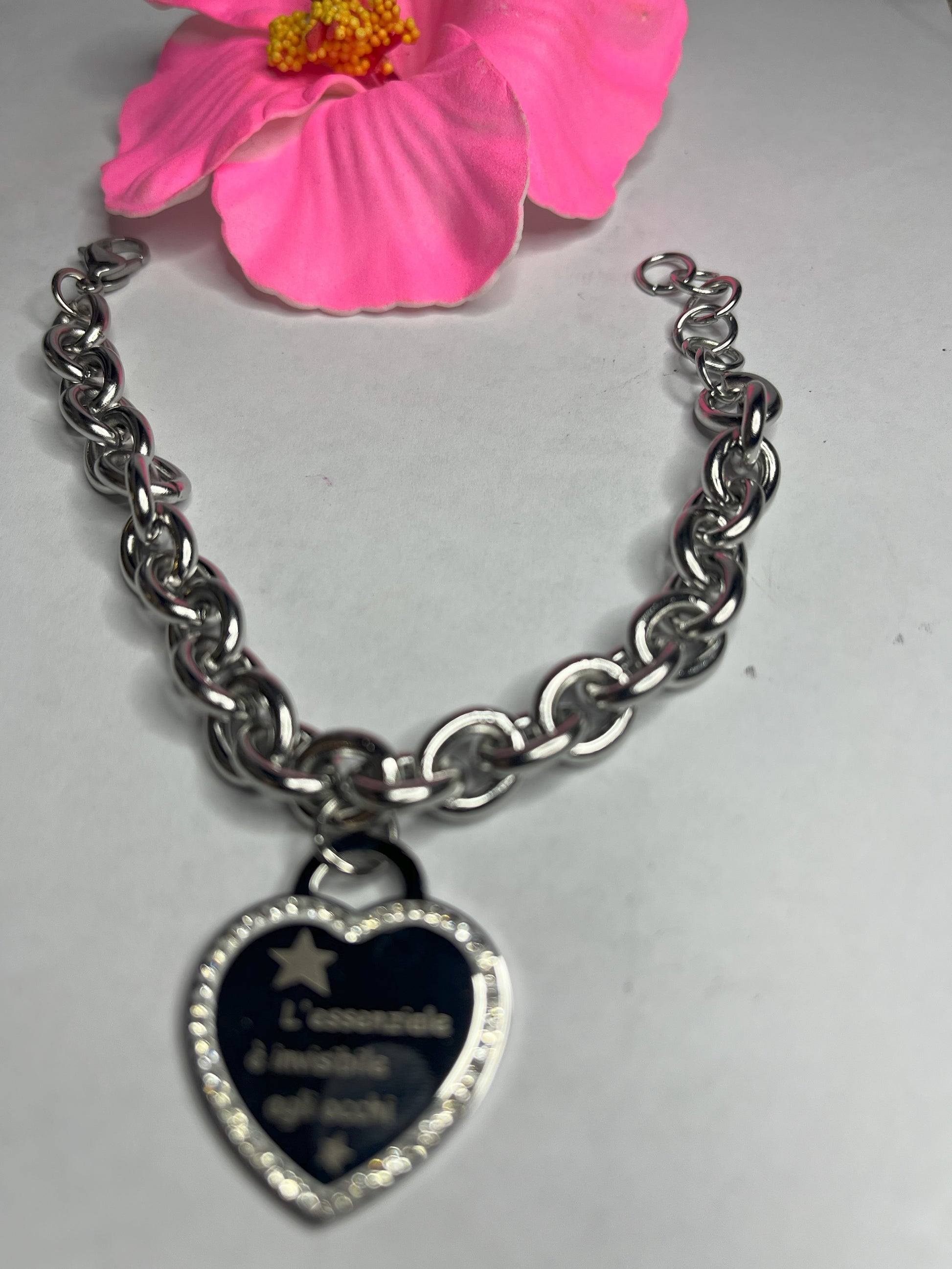 Braccialetto chunky argentato con pendente a cuore nero contenente la scritta "L'essenziale è invisibile agli occhi" su sfondo grigio con petali rosa