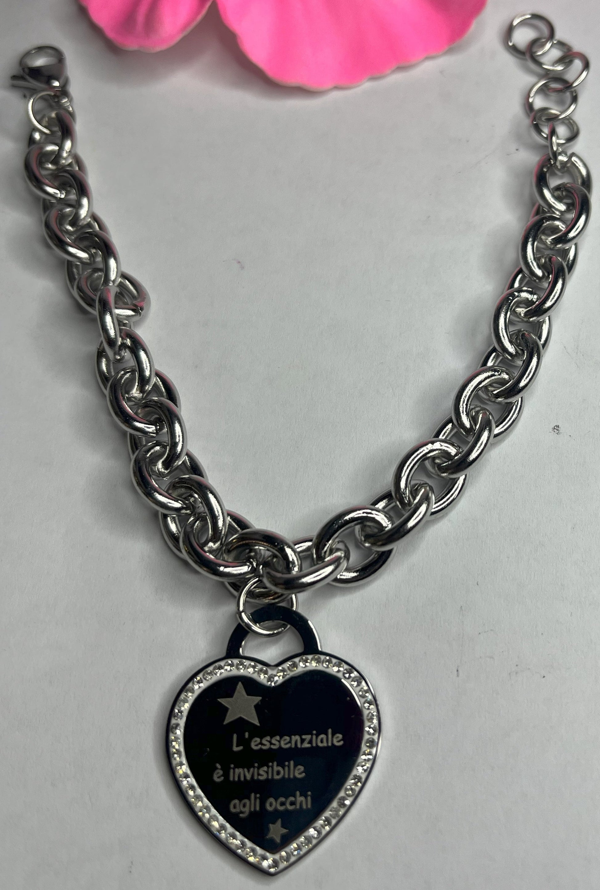 Braccialetto chunky argentato con pendente a cuore nero contenente la scritta "L'essenziale è invisibile agli occhi" su sfondo grigio con petali rosa