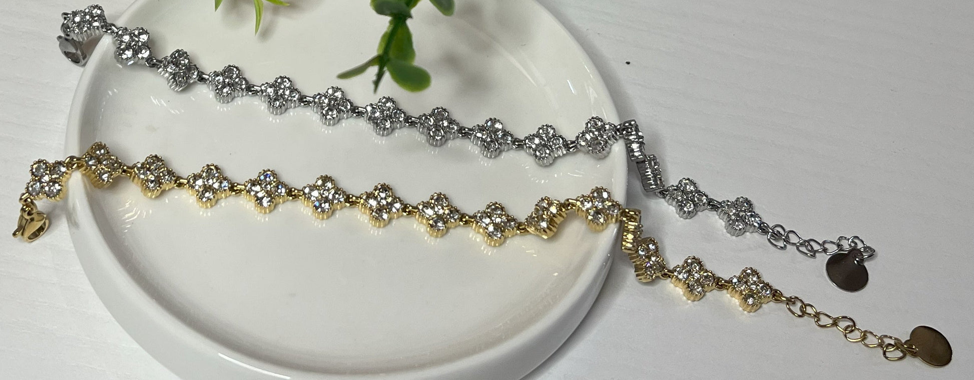 Braccialetti tennis, uno placcato oro, uno placcato argento con quadrifogli portafortuna e cristalli scintillanti su sfondo bianco con foglie verdi