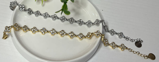 Braccialetti tennis, uno placcato oro, uno placcato argento con quadrifogli portafortuna e cristalli scintillanti su sfondo bianco con foglie verdi