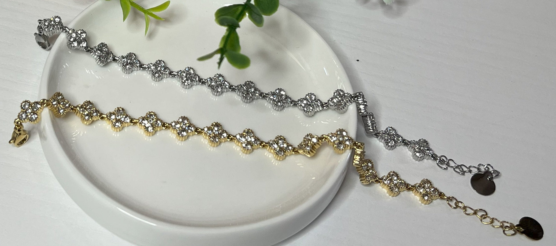 Braccialetti tennis, uno placcato oro, uno placcato argento con quadrifogli portafortuna e cristalli scintillanti su sfondo bianco con foglie verdi