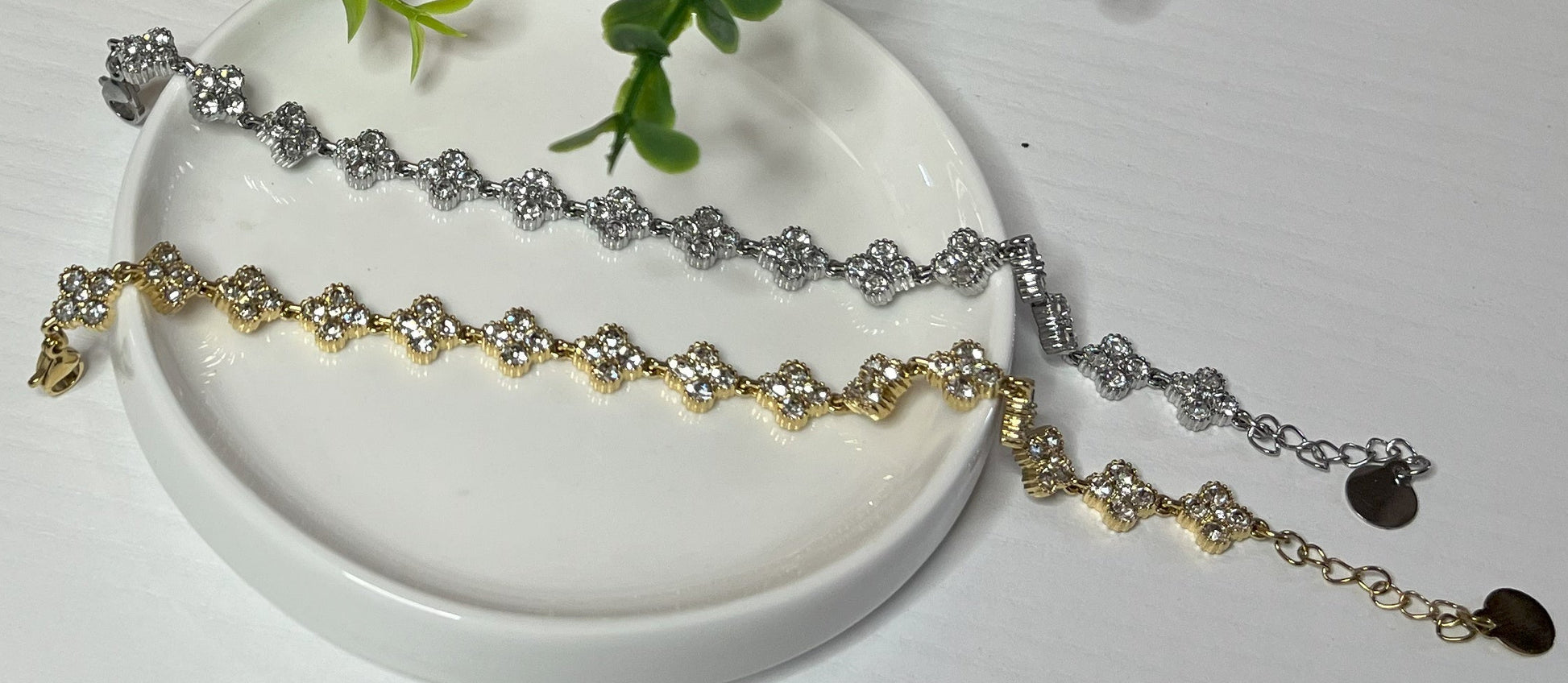 Braccialetti tennis, uno placcato oro, uno placcato argento con quadrifogli portafortuna e cristalli scintillanti su sfondo bianco con foglie verdi