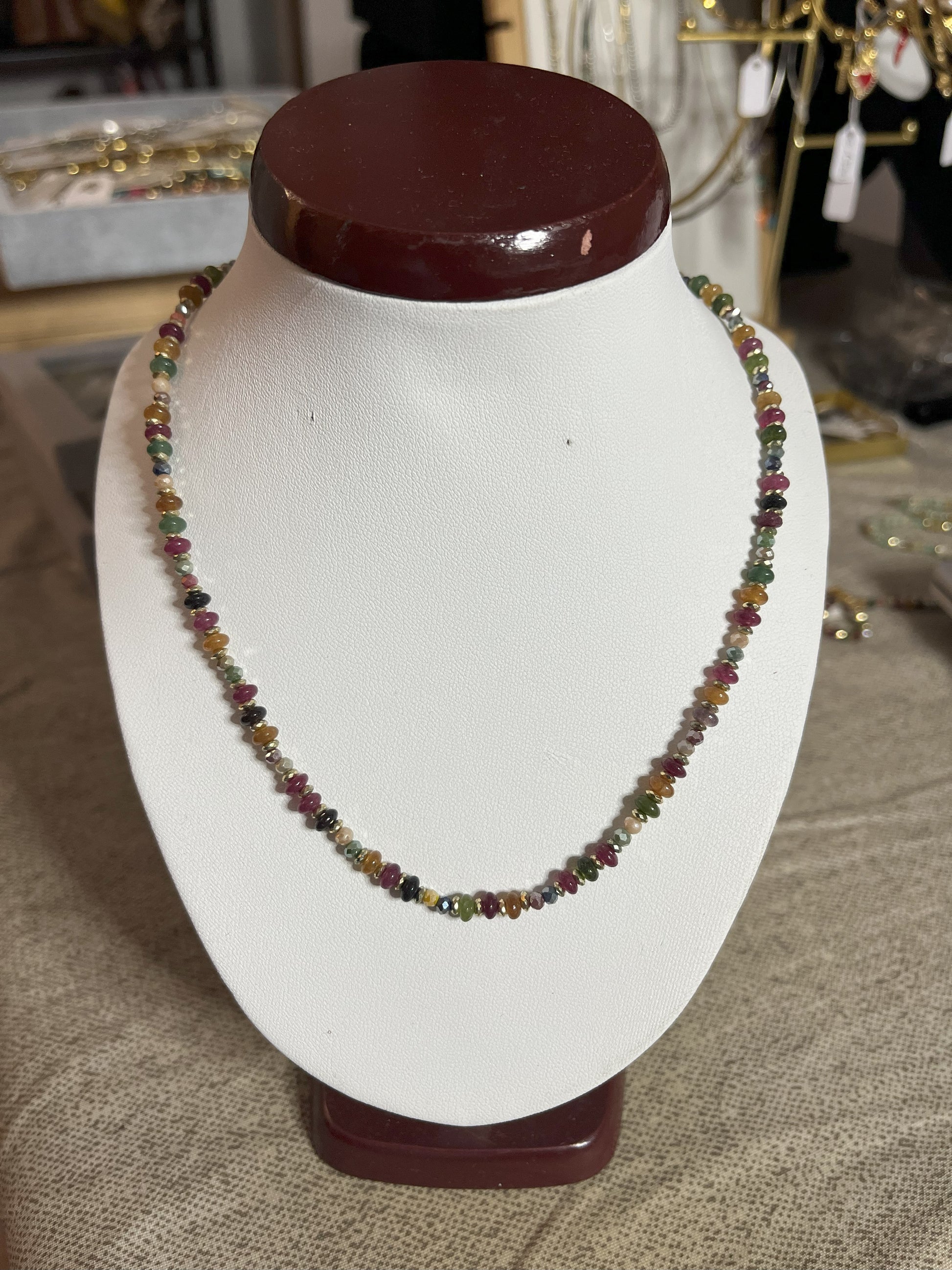 Collana Mosaico Prezioso disponibile in due varianti: multicolore con pietre rosse, verdi, blu e oro, e verde-oro con tonalità smeraldo e dorate, su espositore bianco