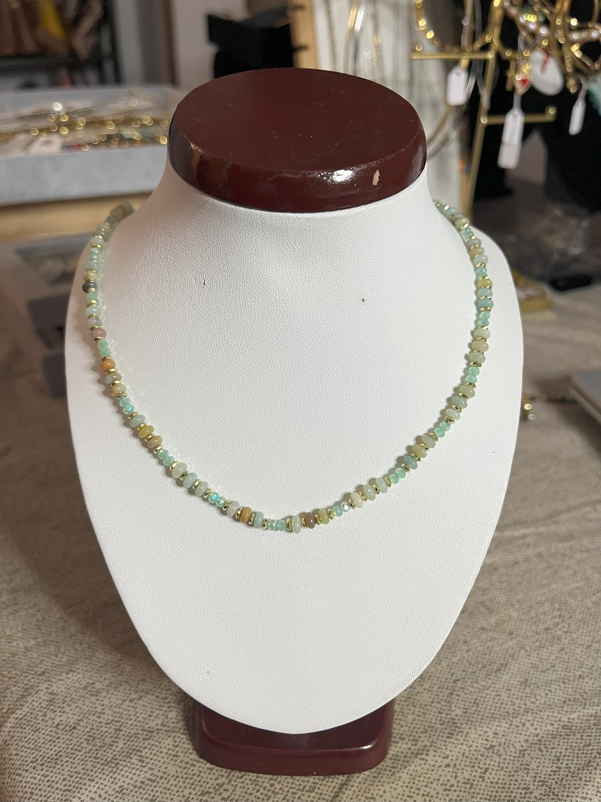 Collana Mosaico Prezioso disponibile in due varianti: multicolore con pietre rosse, verdi, blu e oro, e verde-oro con tonalità smeraldo e dorate, su espositore bianco