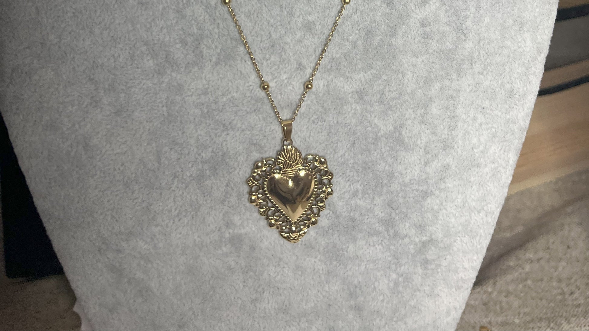 Collana Regina Cuori Sacri in acciaio premium placcato oro con ciondolo a cuore ornamentale barocco e catena decorativa con sfere, esposta su busto grigio