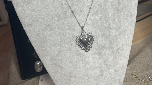 Collana Regina Cuori Sacri in acciaio premium placcato oro con ciondolo a cuore ornamentale barocco e catena decorativa con sfere, esposta su busto grigio