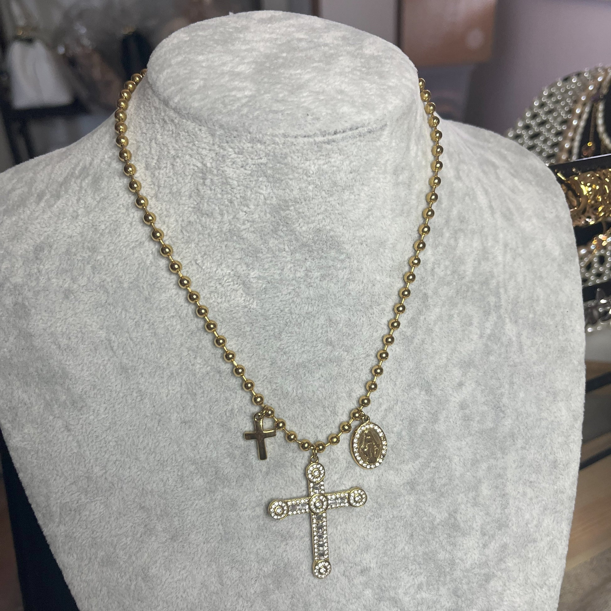 Collana Sfere Croce in acciaio premium placcato oro con catena a sfere e ciondolo croce decorato, esposta su busto grigio