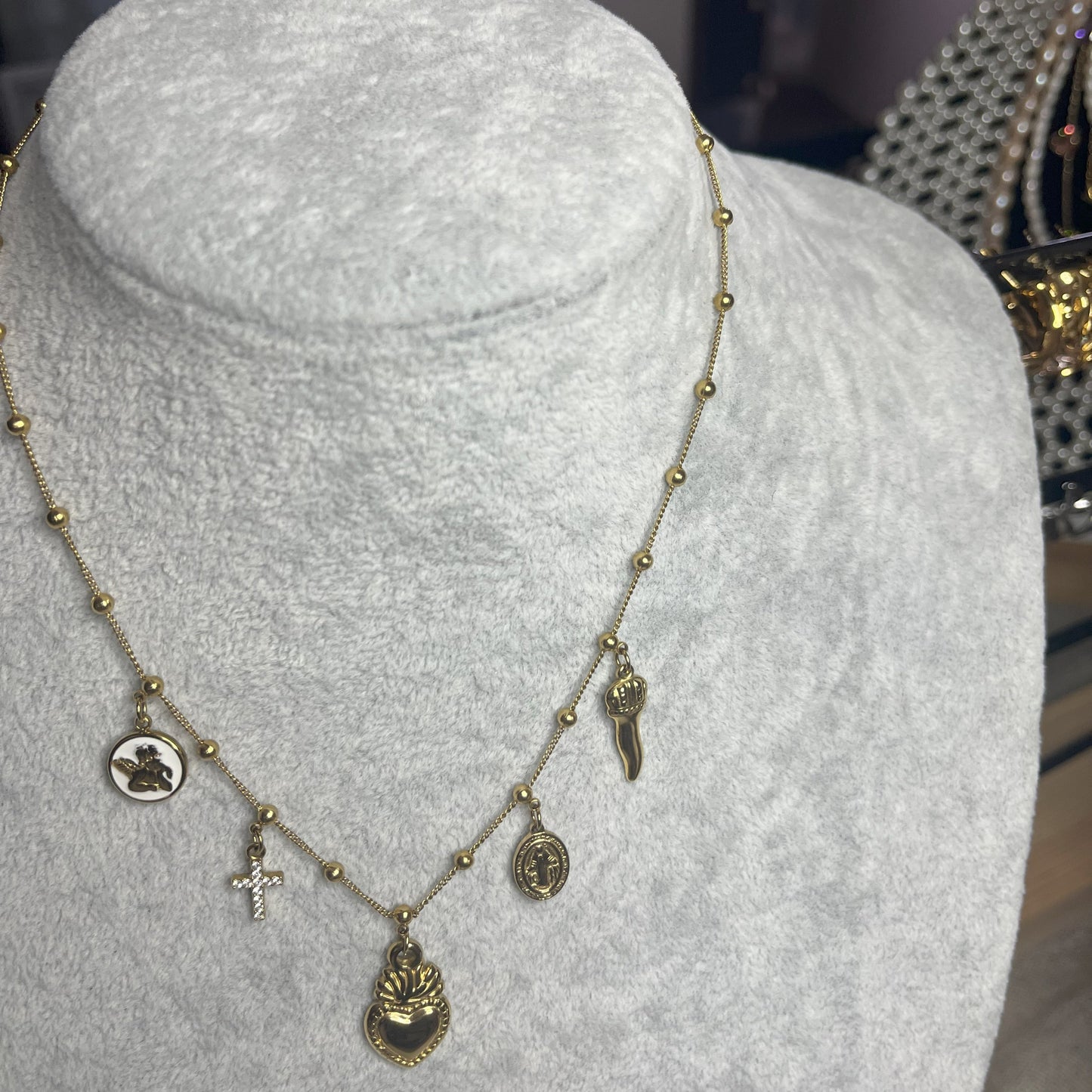 Collana Zoe Charms con Angelo in acciaio premium placcato oro con ciondoli multipli e charm angelo, indossata su busto espositore grigio