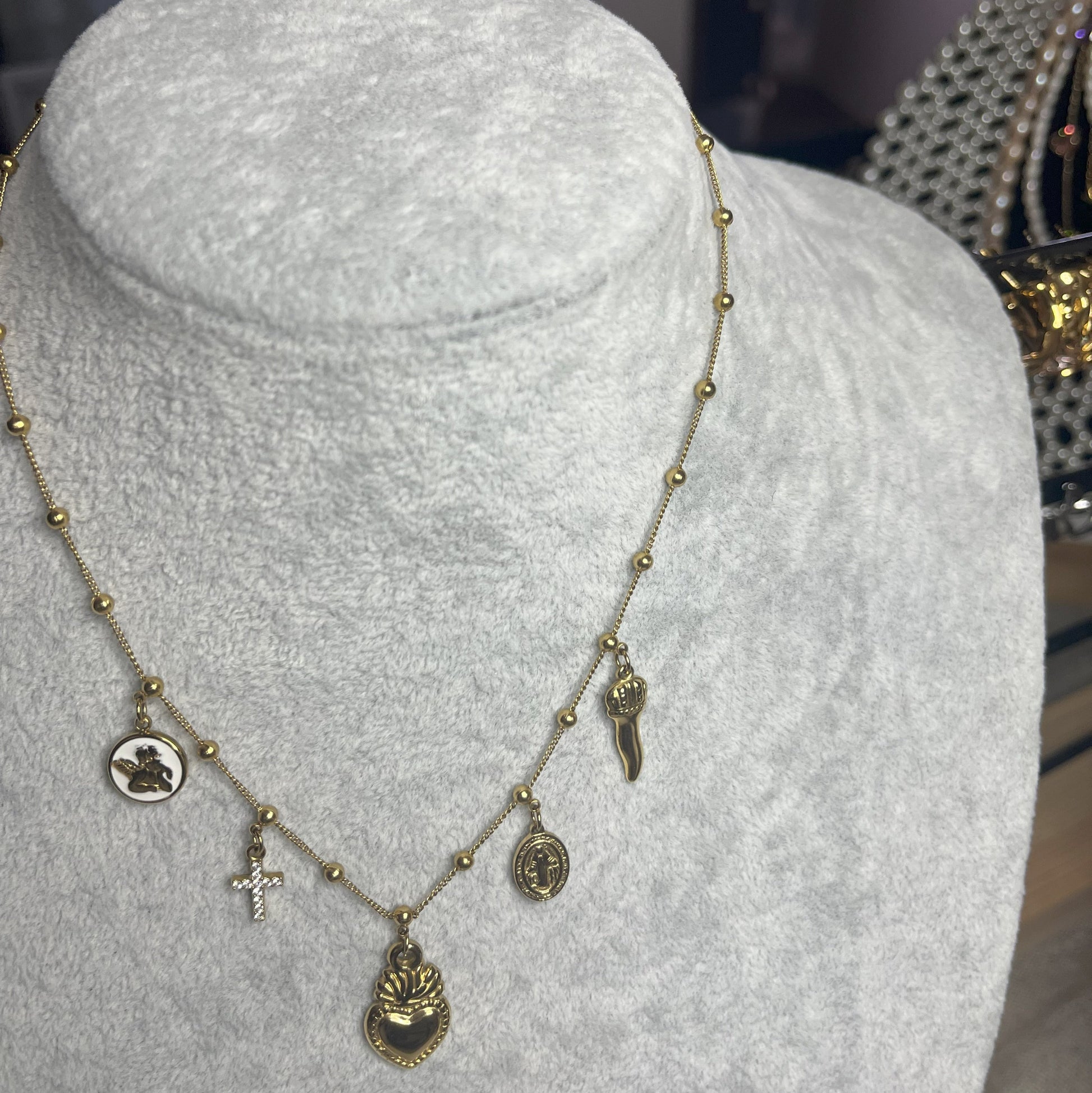 Collana Zoe Charms con Angelo in acciaio premium placcato oro con ciondoli multipli e charm angelo, indossata su busto espositore grigio