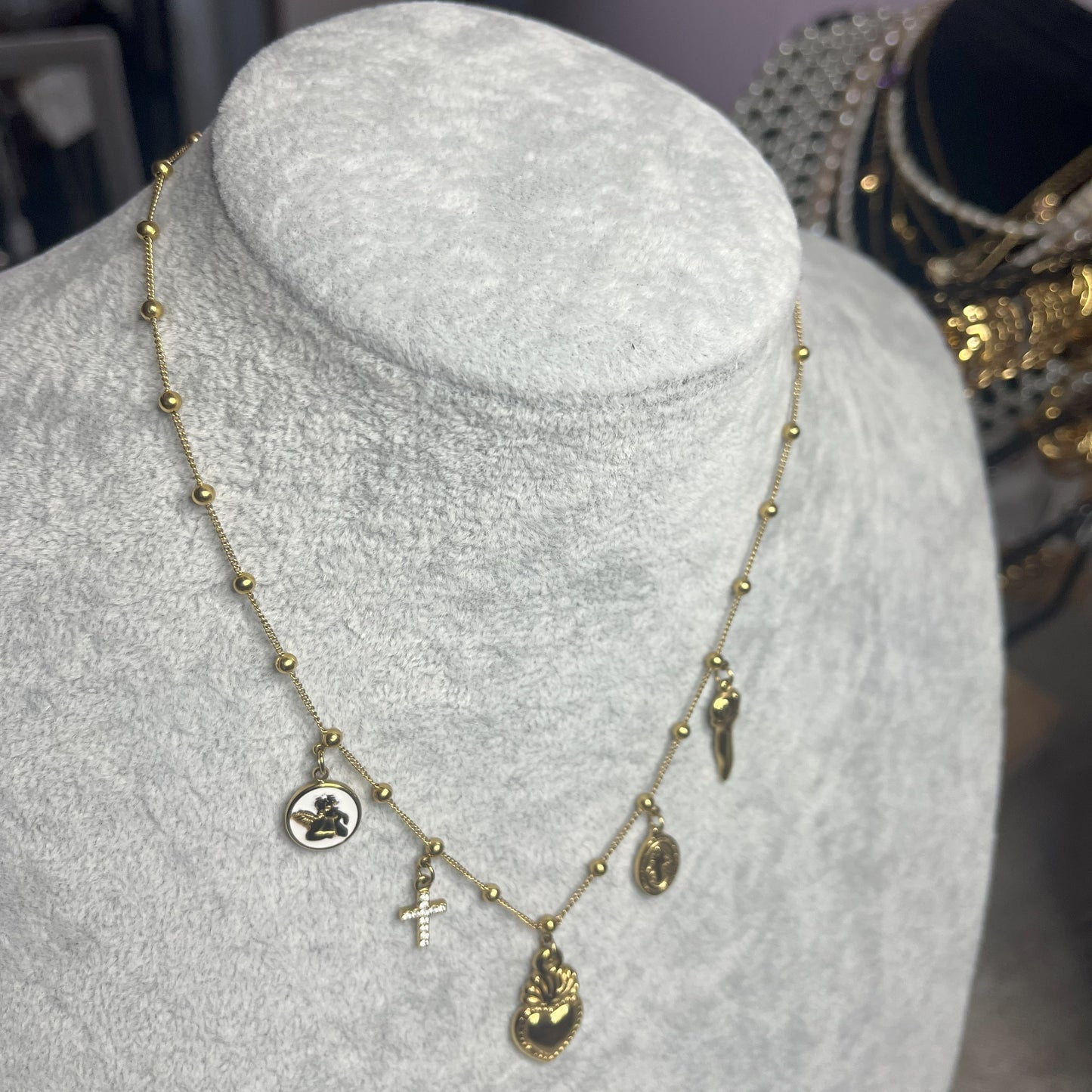 Collana Zoe Charms con Angelo in acciaio premium placcato oro con ciondoli multipli e charm angelo, indossata su busto espositore grigio