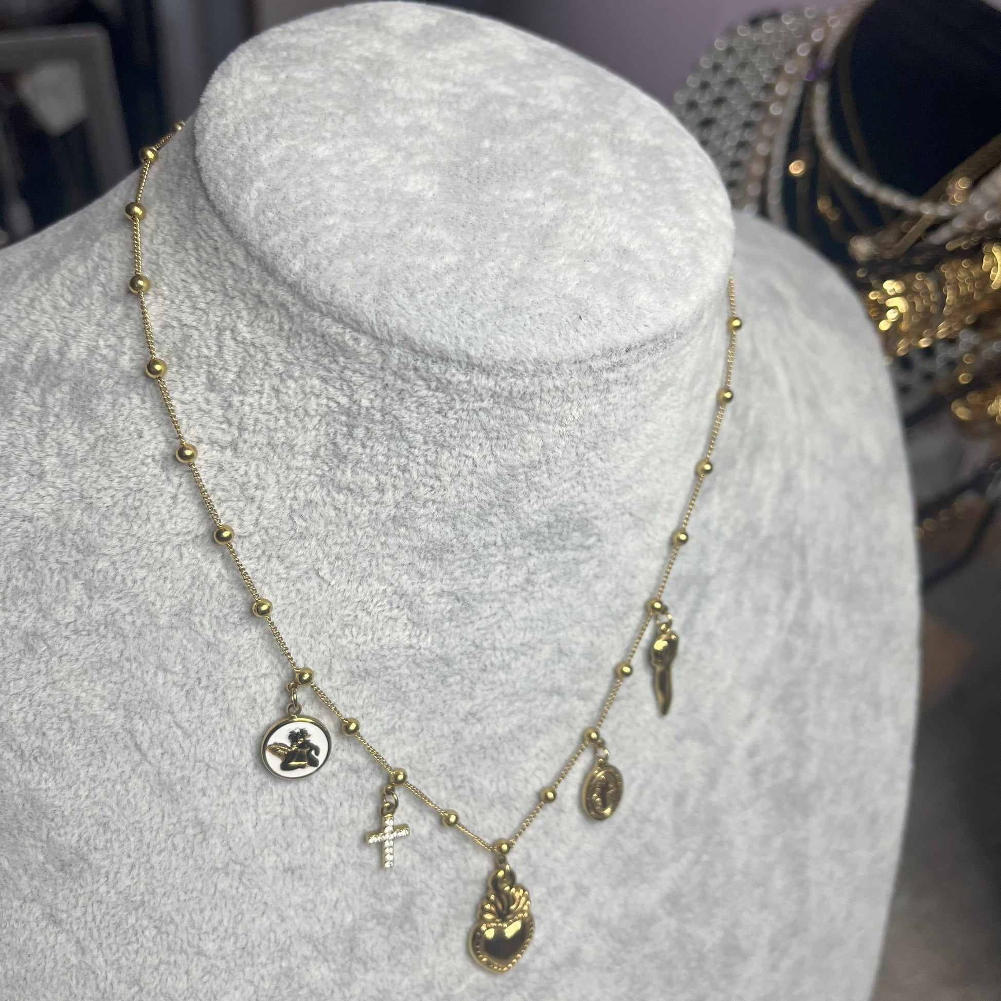 Collana Zoe Charms con Angelo in acciaio premium placcato oro con ciondoli multipli e charm angelo, indossata su busto espositore grigio